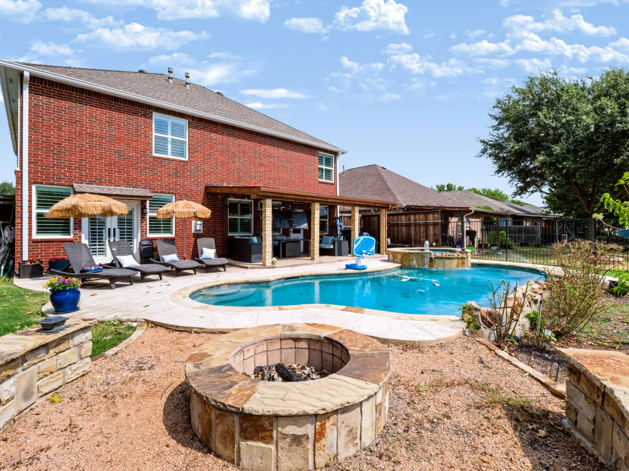 Brick exterior with a covered patio, an in-ground swimming pool, a hot tub with a waterfall feature, and a stone-clad fire pit