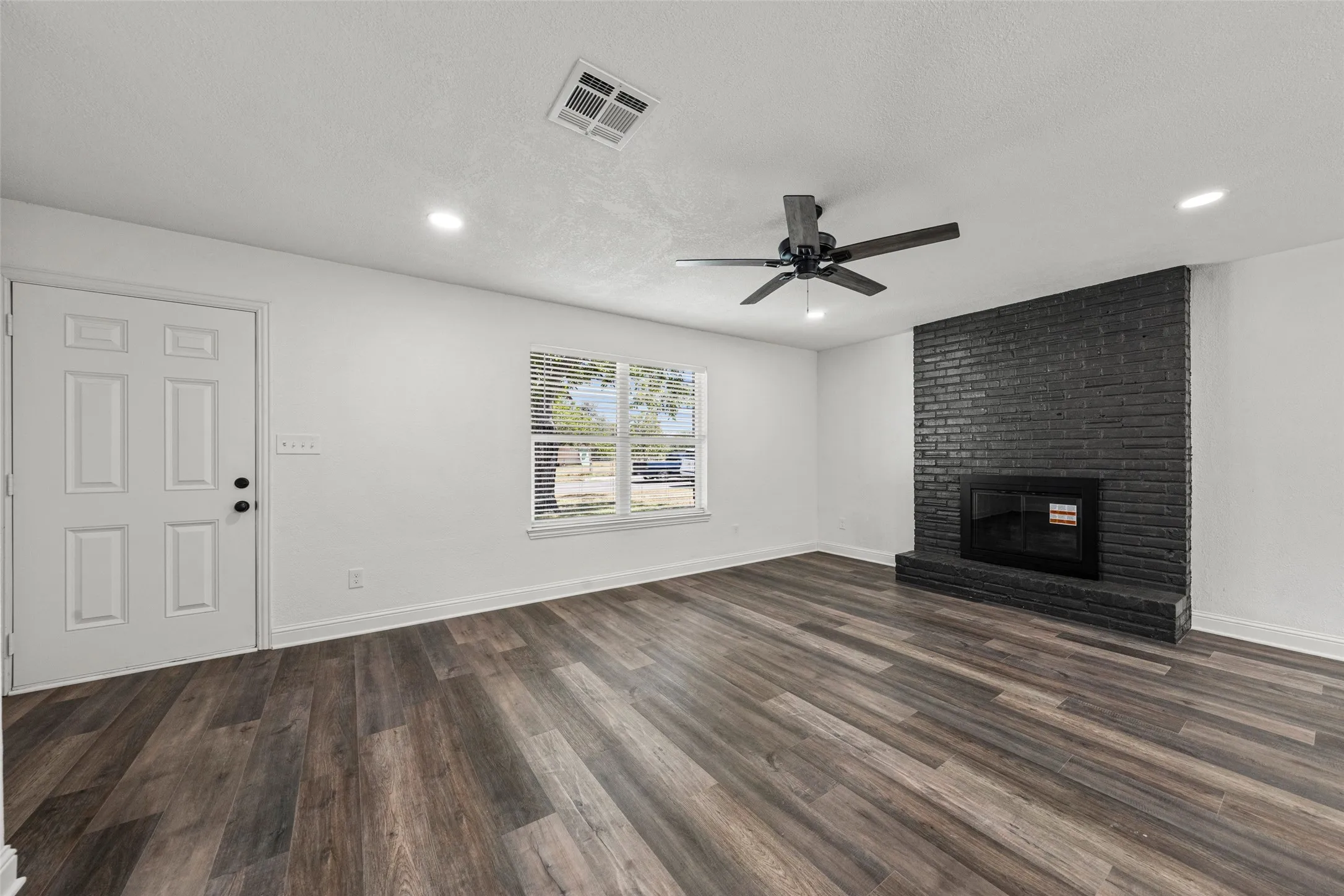Wood-finish flooring throughout, featuring a painted brick fireplace with a hearth, a ceiling fan with integrated lighting, and recessed ceiling lights