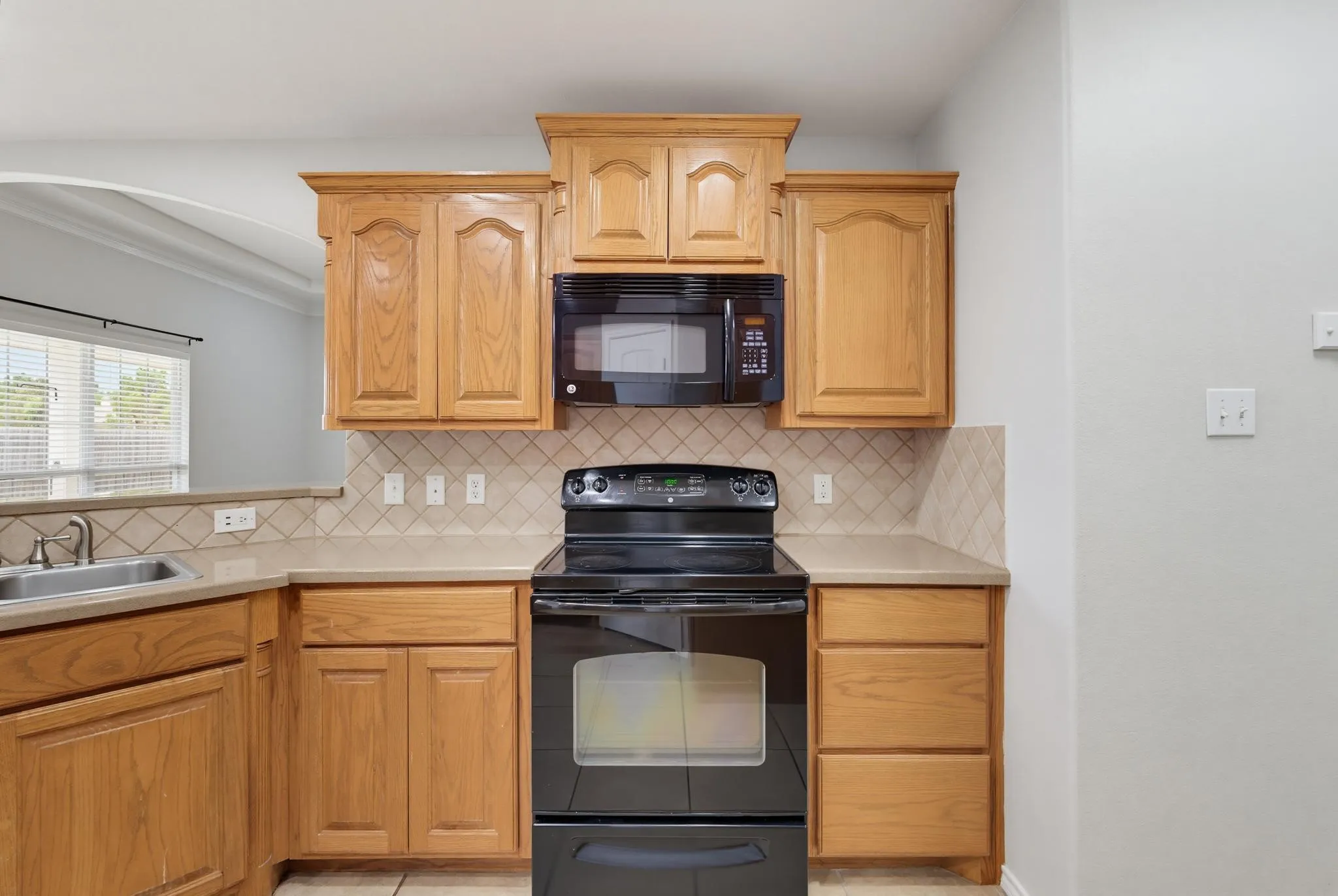 Efficient kitchen featuring warm wood cabinetry with raised-panel doors, black electric range with window, tile backsplash, and stainless steel sink