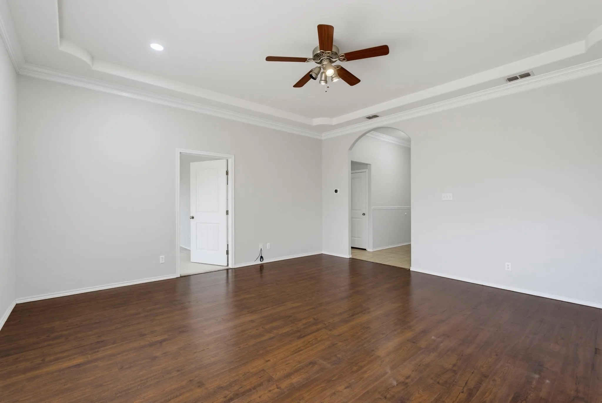 Wood-finish flooring, tray ceiling with crown molding, ceiling fan, recessed lighting, and arched doorway