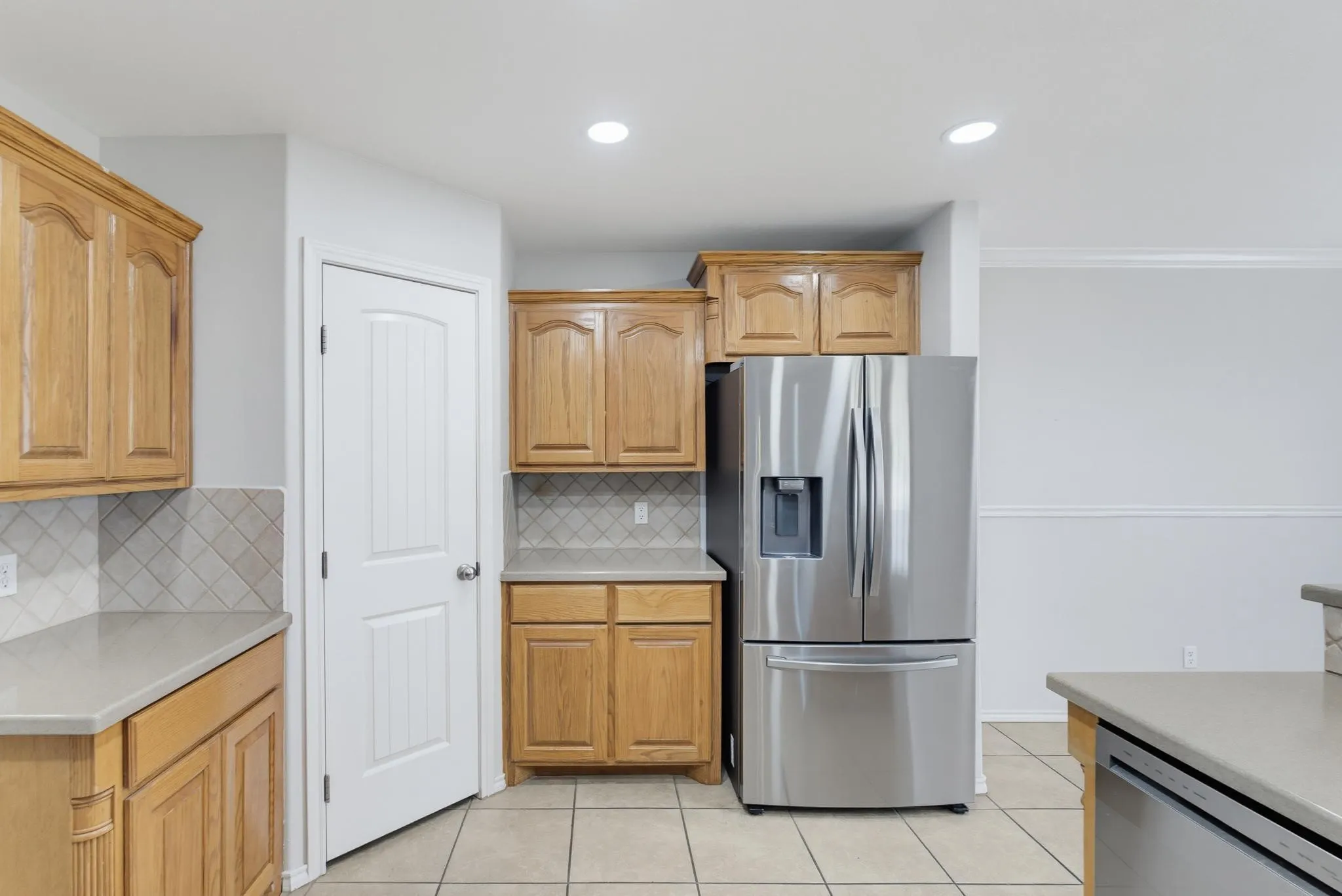 Bright kitchen featuring natural wood cabinetry, stainless steel French-door refrigerator with ice and water dispenser, ceramic tile flooring, recessed lighting, and a geometric tile backsplash