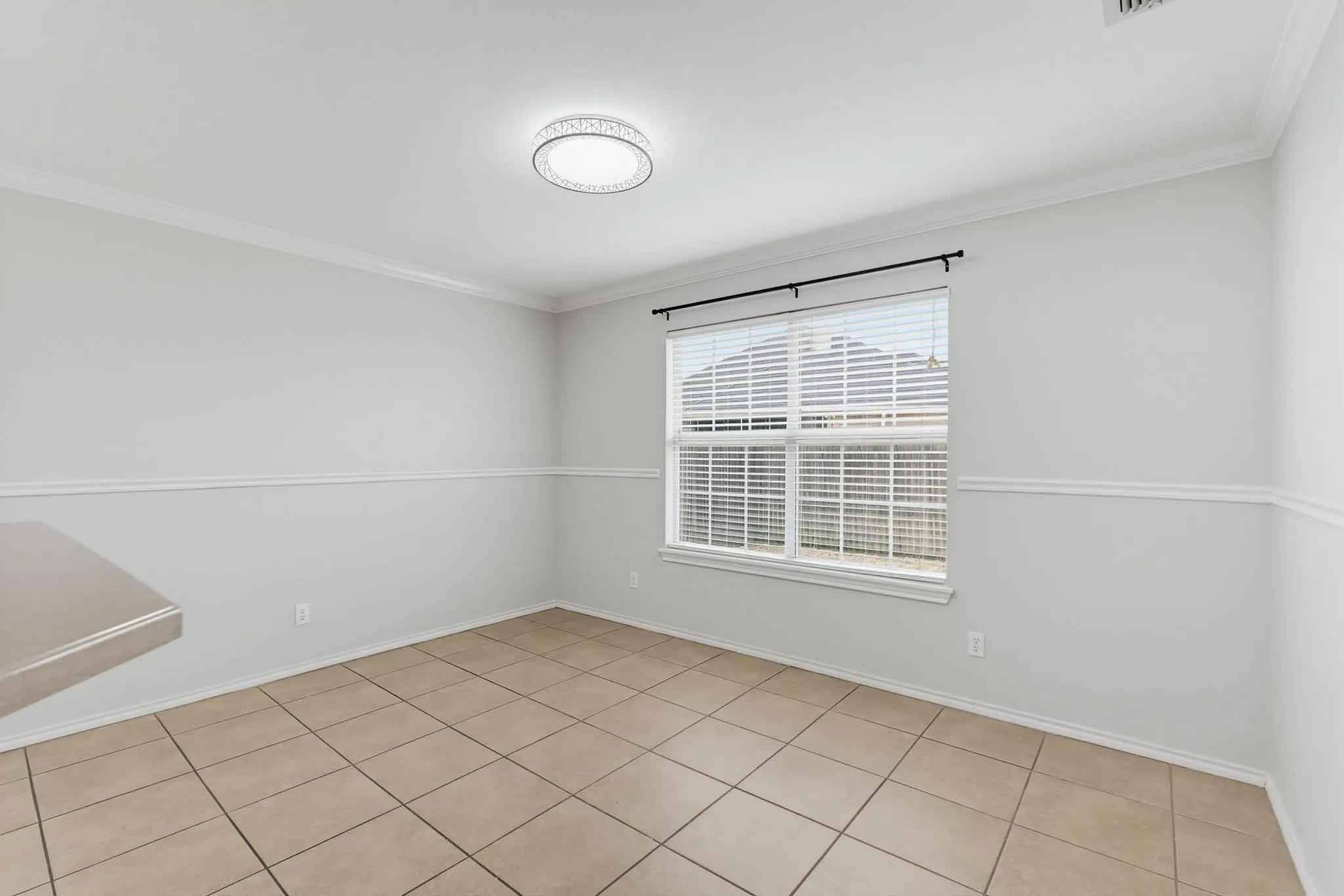 Bright secondary bedroom featuring neutral wall finishes with chair rail detailing, ceramic tile flooring throughout, and a large window with plantation shutters and black hardware rod for natural light and ventilation