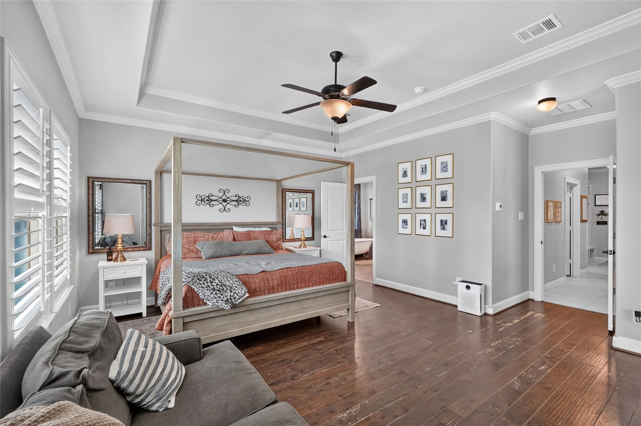 Spacious primary bedroom suite featuring a four-poster canopy bed with wood frame, warm wood-finish flooring throughout, coffered ceiling with recessed lighting, and ceiling fan with integrated light fixture