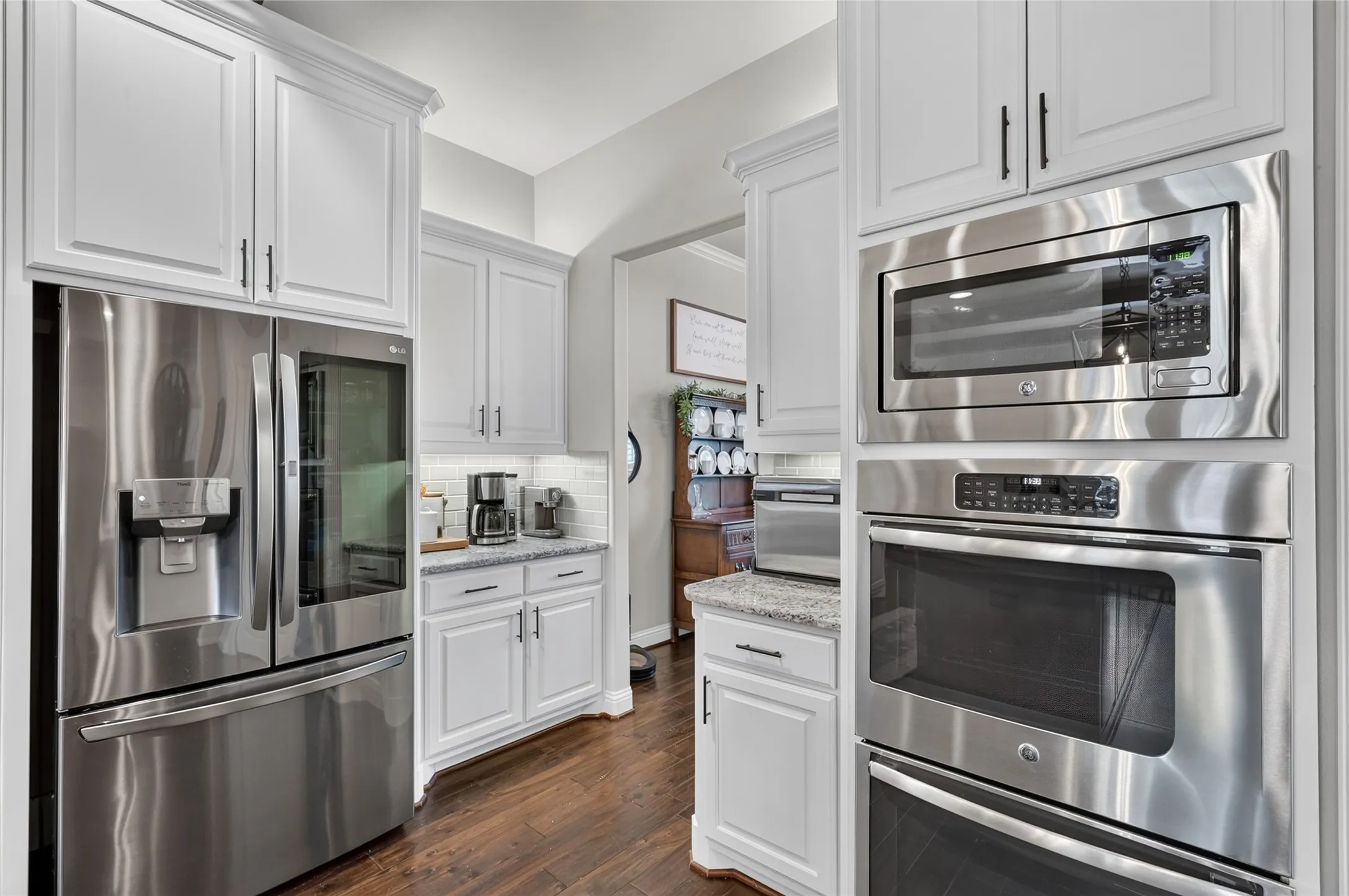 Chef's kitchen featuring stainless steel French-door refrigerator, dual wall-mounted ovens with microwave, gas cooktop, white shaker cabinetry with hardware accents, and wood-finish flooring throughout