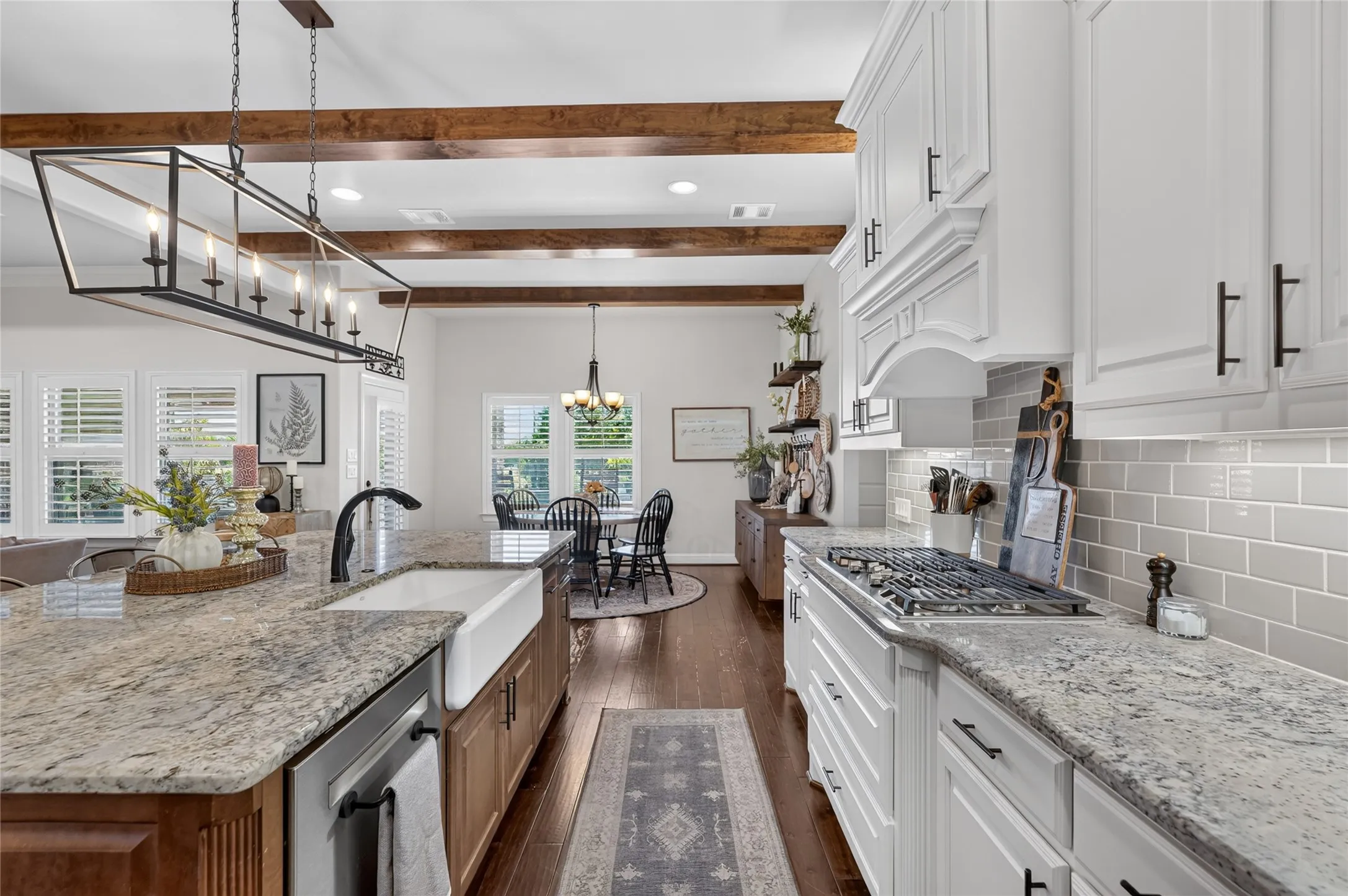Chef's kitchen featuring exposed wood beam ceiling, dual islands with stone countertops and undermount sink, gas cooktop with stainless steel appliances, white cabinetry with hardware accents, subway tile backsplash, and hardwood flooring throughout the open-concept space