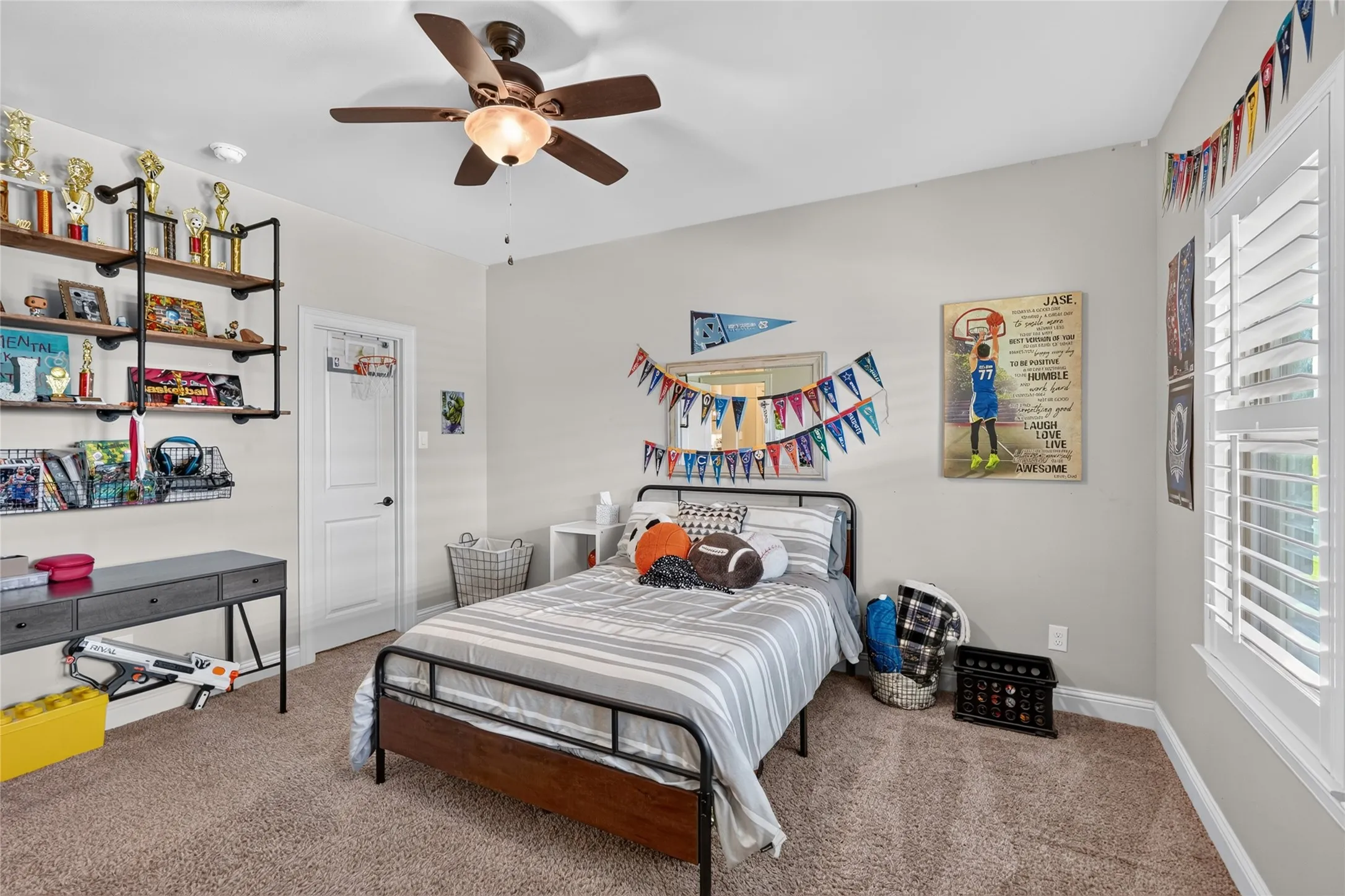 Ceiling fan with integrated lighting, window with plantation shutters, wall-mounted open shelving, light-toned carpeting, and a white interior door