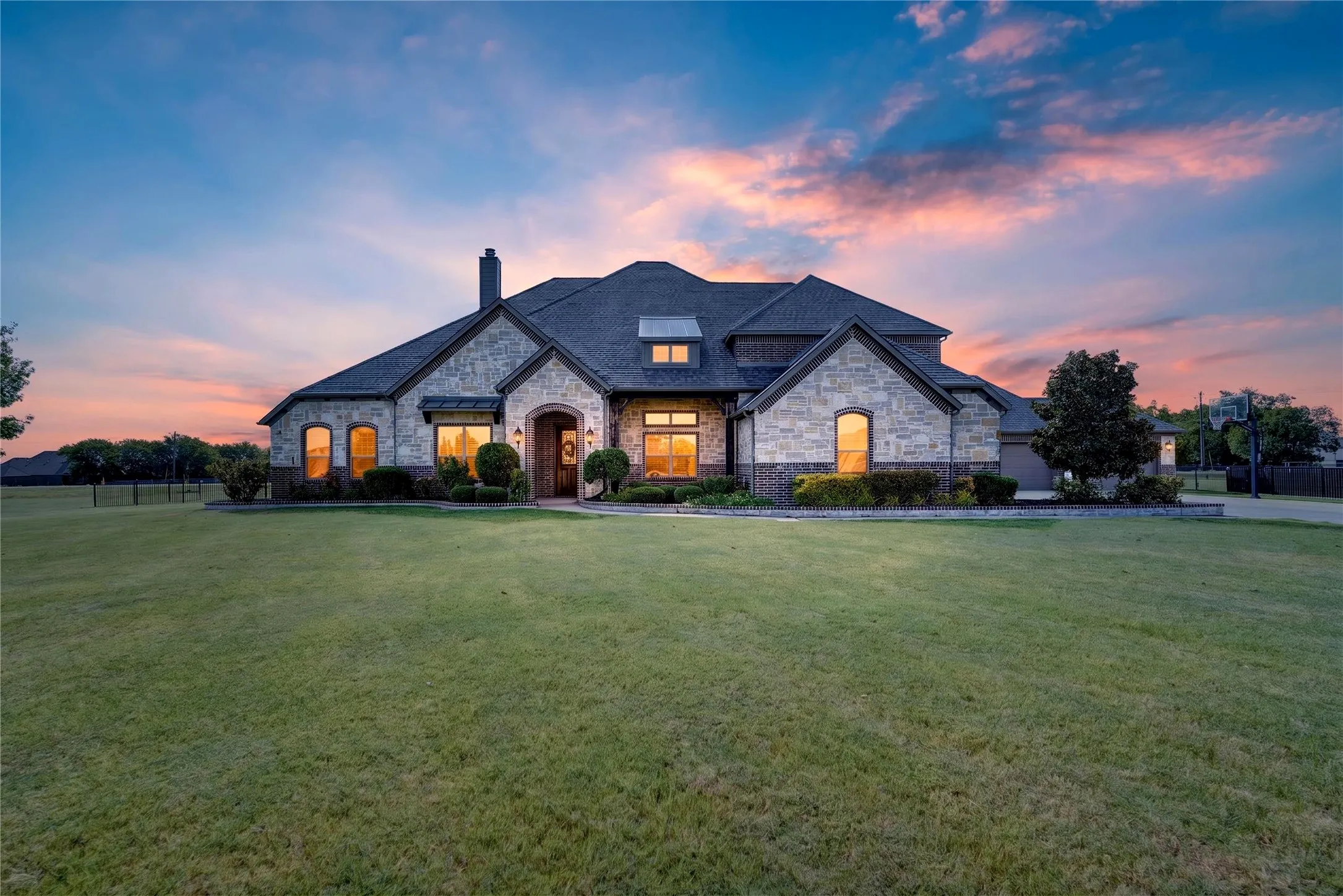 Striking two-story residence featuring a stone and brick exterior, multiple gabled rooflines with dark asphalt shingles, and abundant windows with warm interior lighting