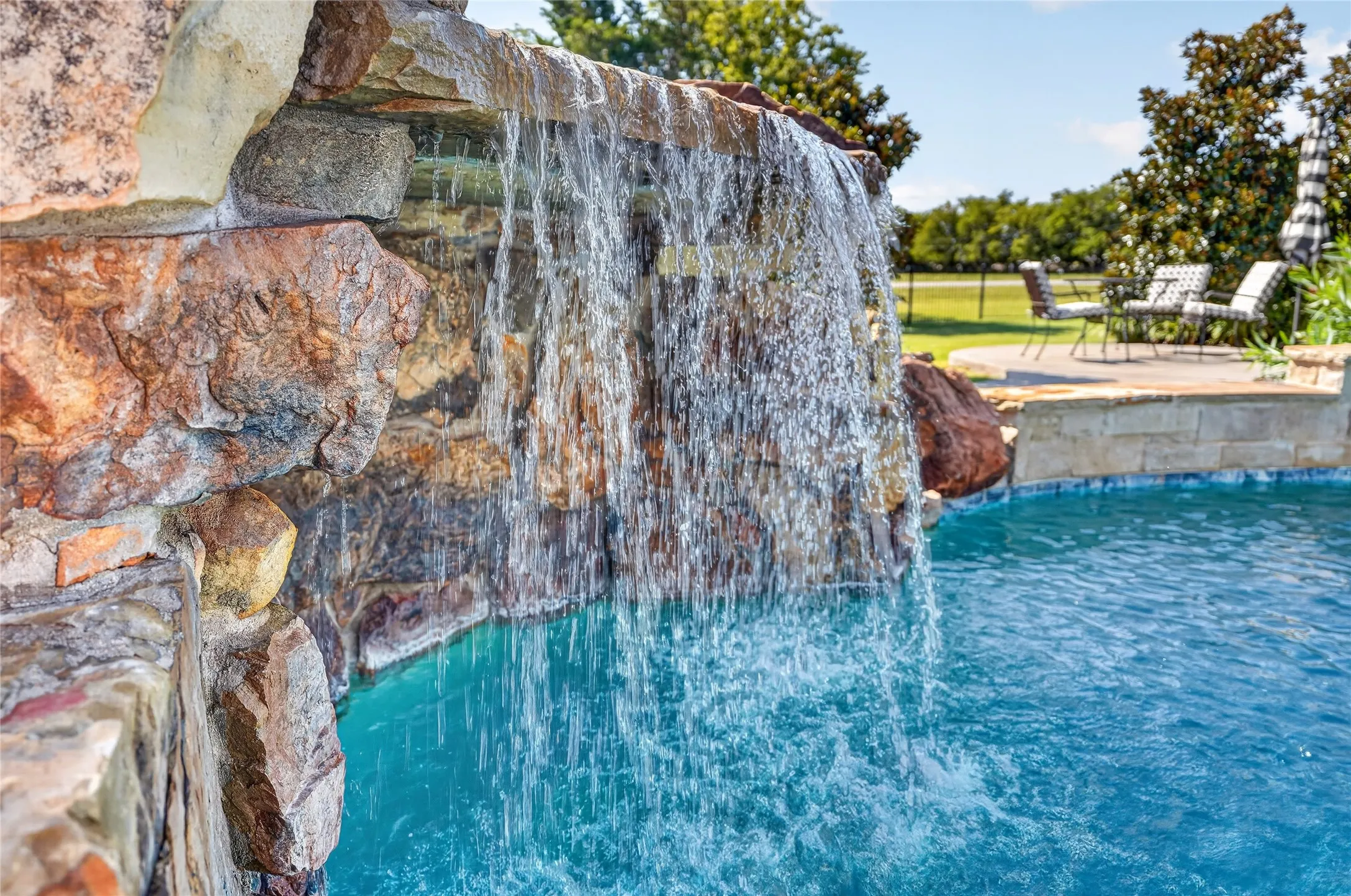 Sparkling resort-style swimming pool featuring a cascading waterfall feature and turquoise water, bordered by natural stone accents and mature landscaping with lush greenery visible beyond the property perimeter