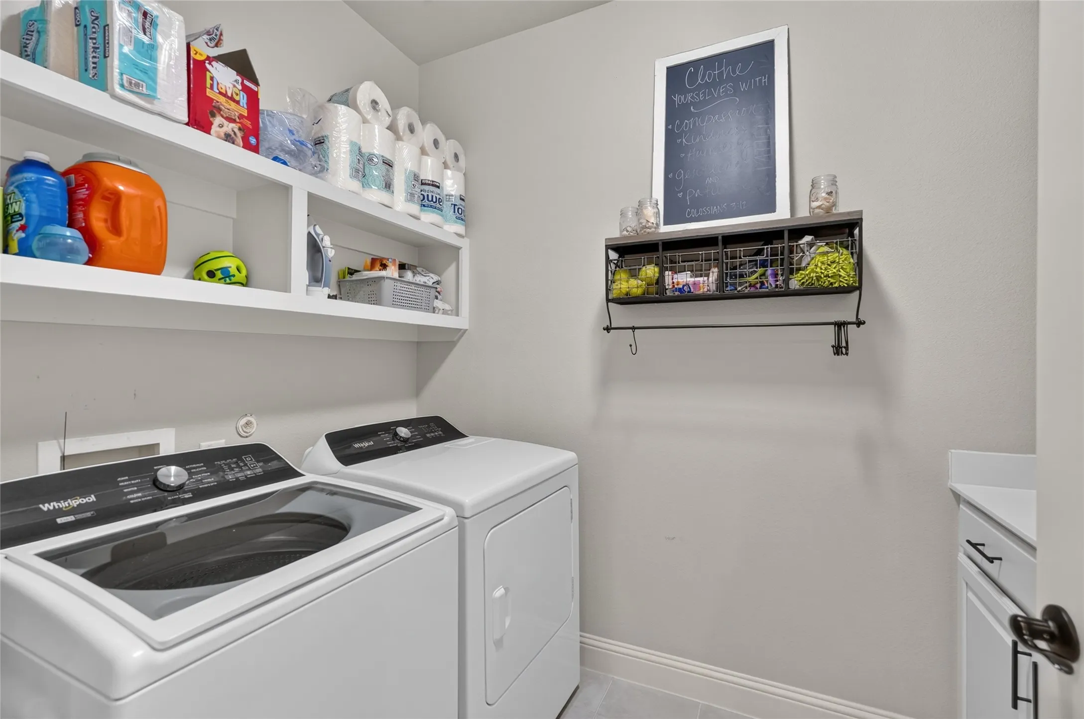 Dedicated laundry room featuring side-by-side washer and dryer with modern white finish, open shelving with storage containers, chalkboard-framed wall organizer, and neutral wall finishes ideal for efficient household management
