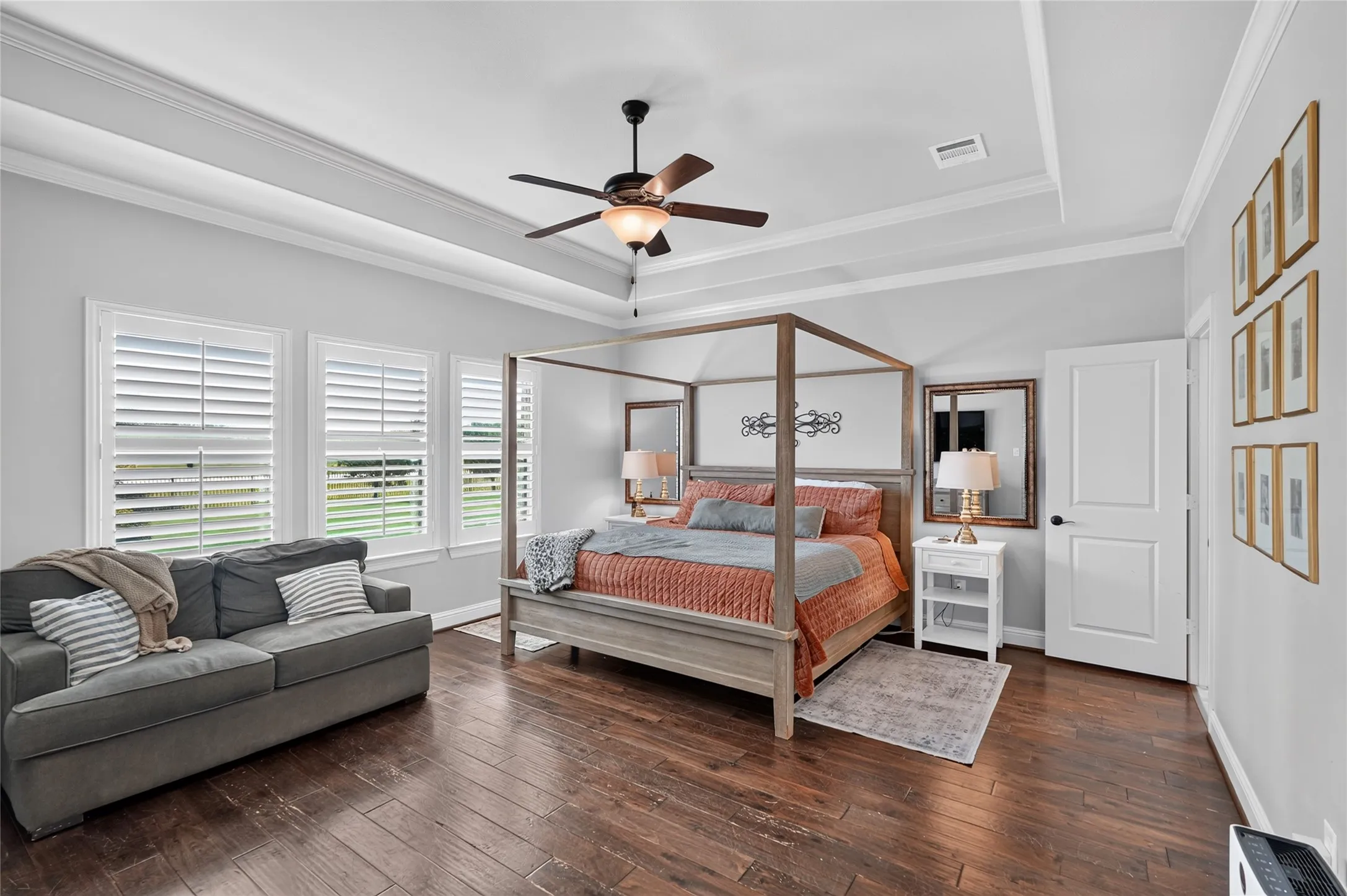 Spacious primary bedroom suite featuring a four-poster bed frame, wood-finish hardwood flooring, coffered ceiling with ceiling fan, plantation shutters along expansive windows, ensuite bathroom with vanity, and a comfortable seating area with built-in sofa