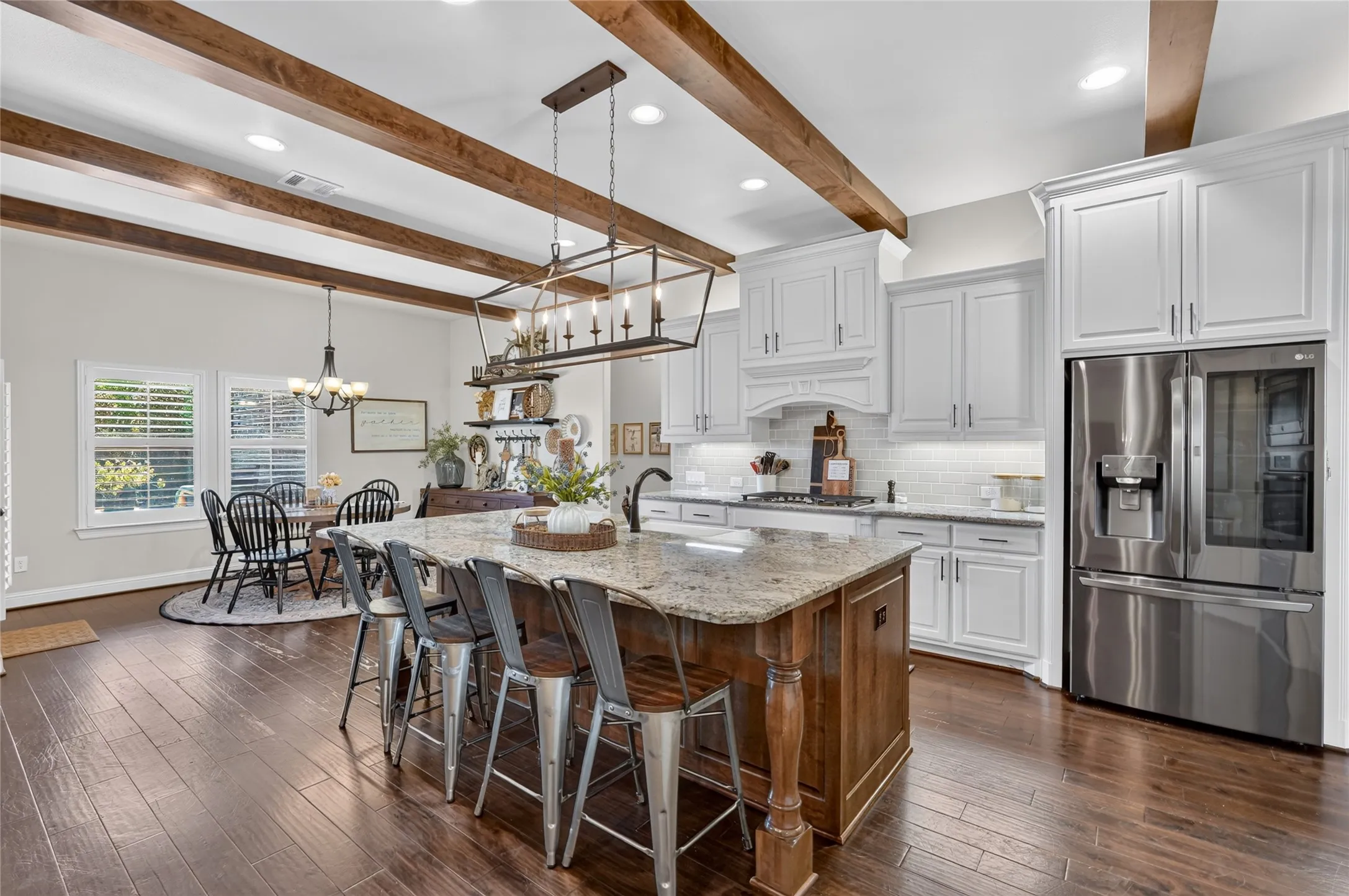 Gourmet kitchen featuring white cabinetry with shaker-style doors, a two-tier island with wood-finish base and stone countertops, stainless steel French-door refrigerator, exposed wood beam ceiling details, hardwood flooring, and pendant lighting over the island workspace