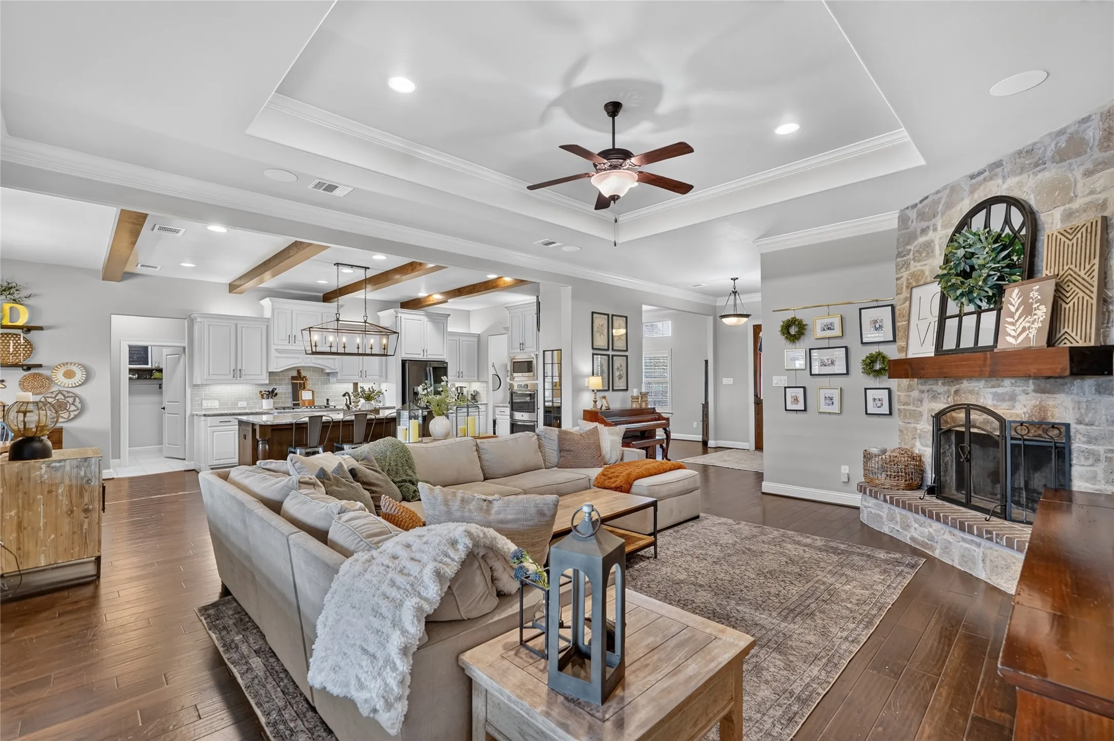 Wood-finish flooring, stone-clad fireplace with wood mantle, tray ceiling with ceiling fan, exposed wood beams, and a kitchen with white cabinetry