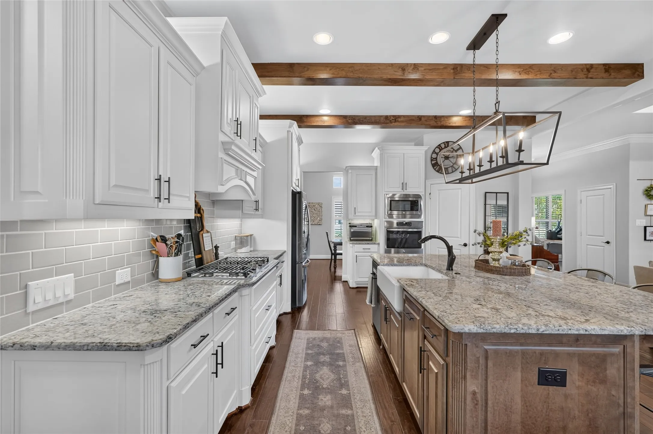 White shaker cabinetry with dark hardware, light-toned granite countertops, wood-finish flooring, a farmhouse sink, and exposed ceiling beams