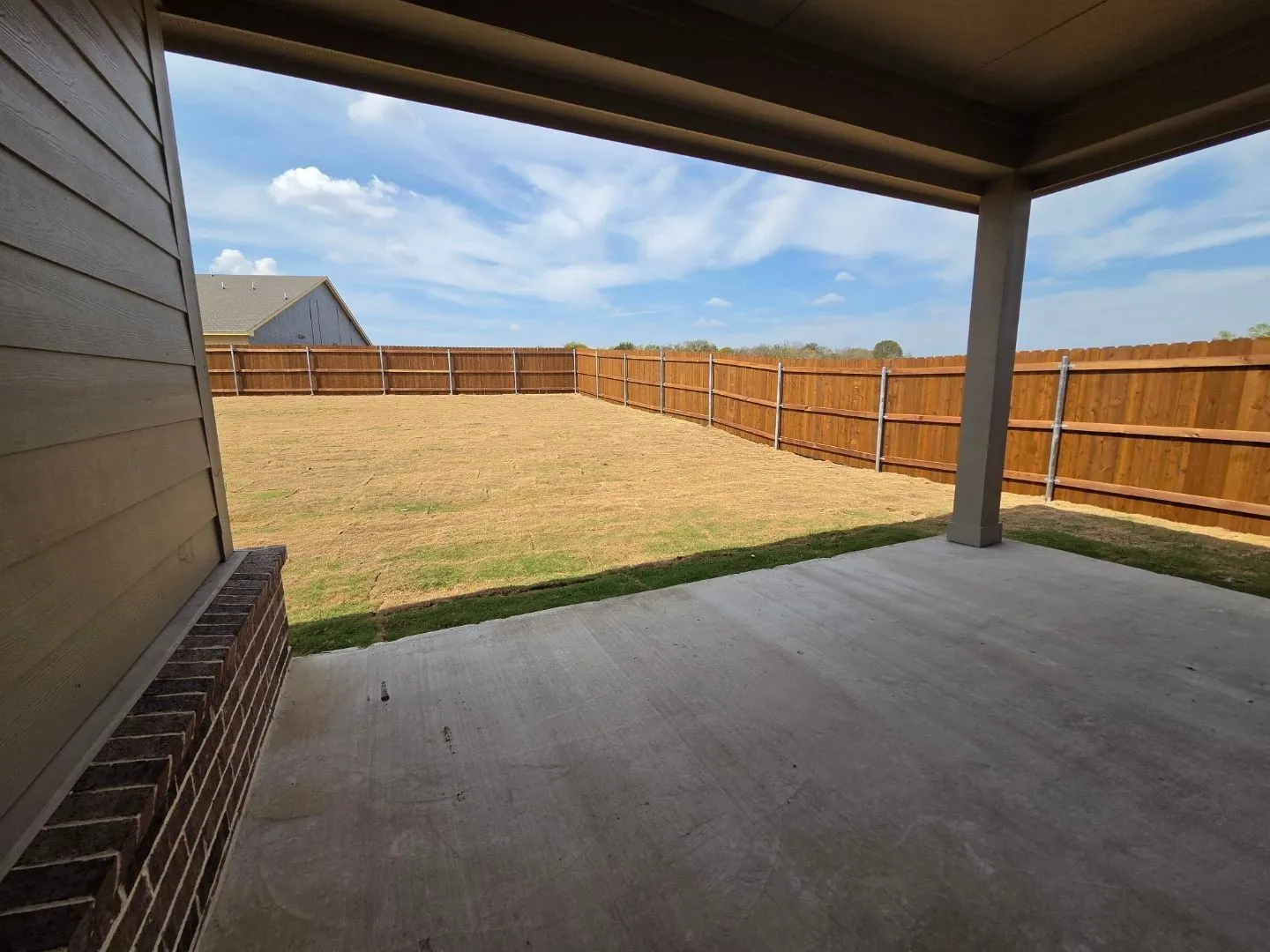 Covered concrete patio with a wood privacy fence, expansive lawn area, and a brick exterior accent