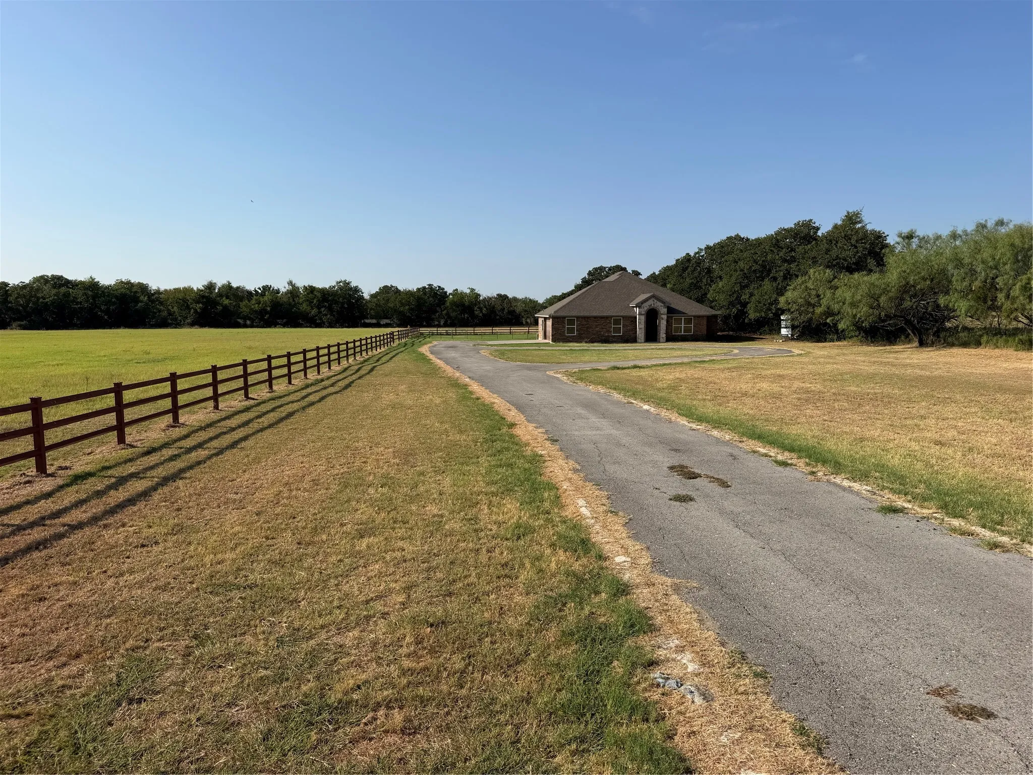 Expansive rural property featuring a long paved driveway lined with black board fencing, mature tree line, and a single-story outbuilding with brick chimney situated on manicured grounds