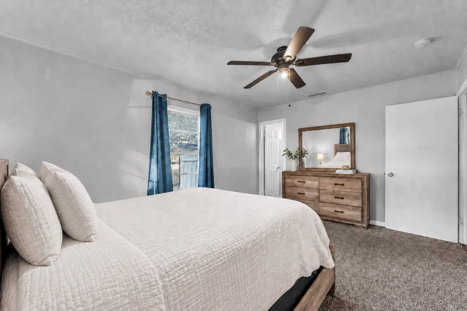 Bright secondary bedroom with ceiling fan, carpeted flooring, wood-finish dresser with mirror, and blue drapery framing the window