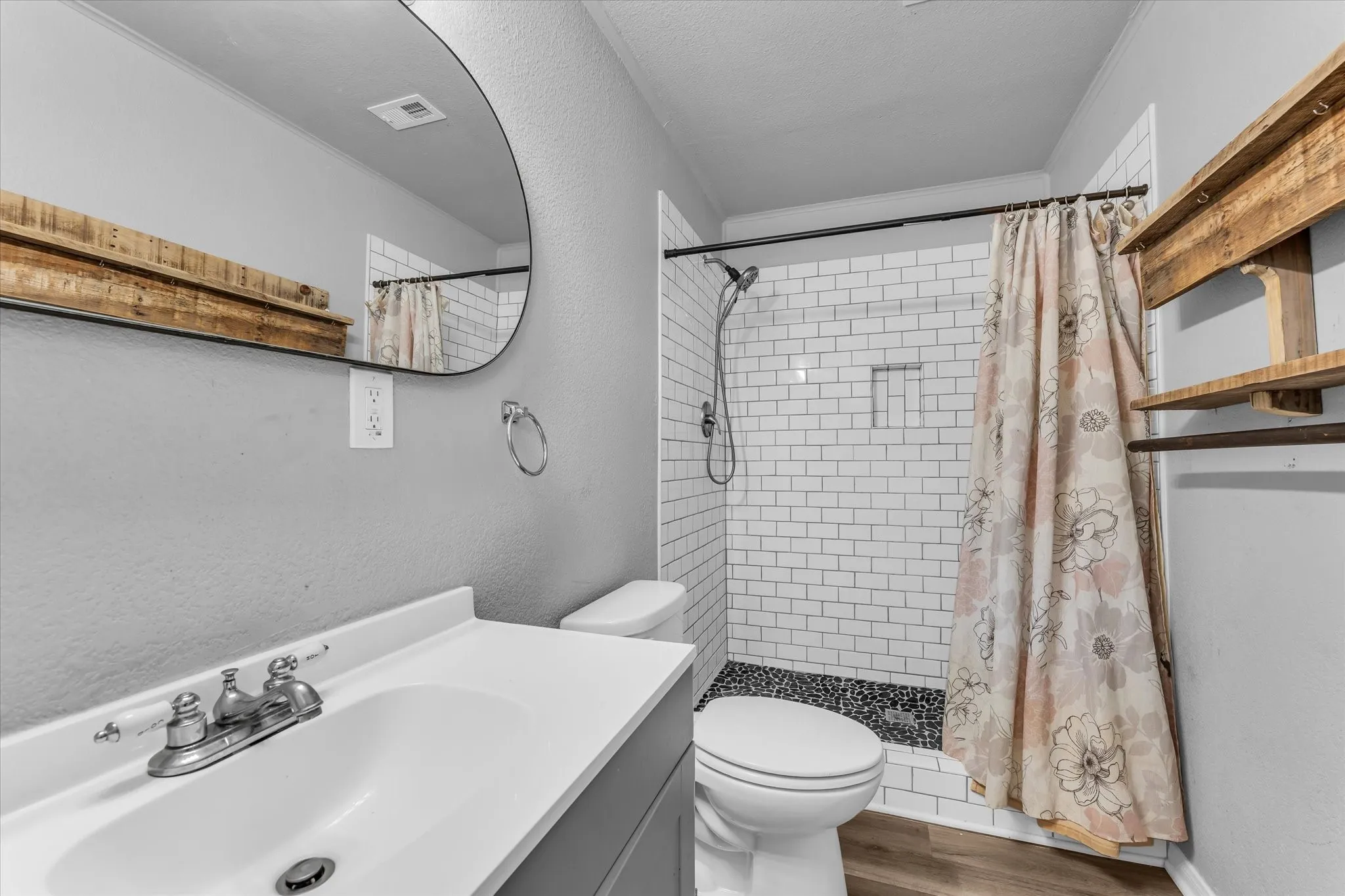 Bathroom featuring a single vanity, round mirror, wood-finish flooring, subway tile shower surround, and black pebble shower pan