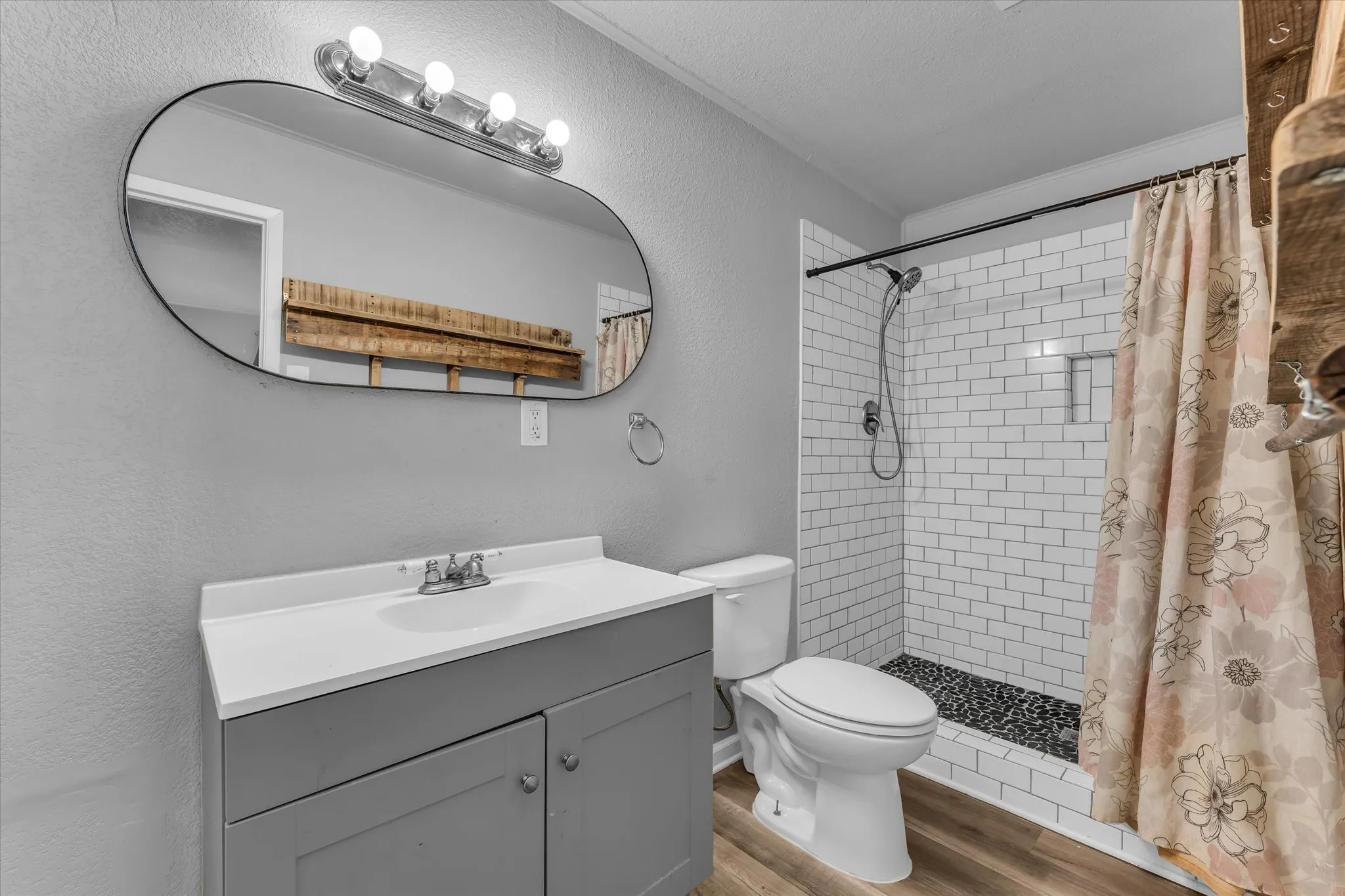 Bathroom featuring a single vanity with a white countertop, oval mirror, walk-in shower with subway tile, pebble flooring, and wood-finish flooring