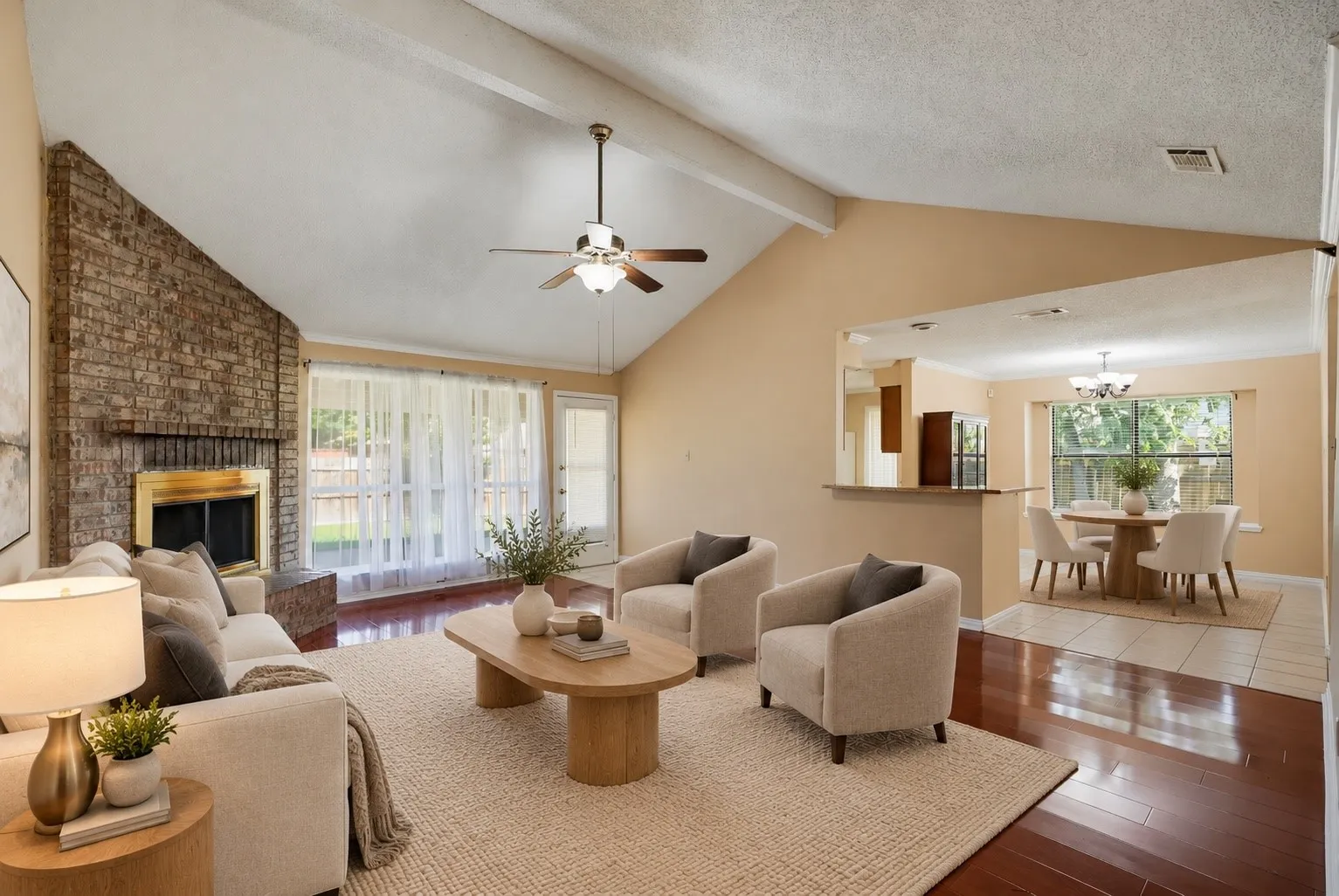Vaulted ceiling with exposed beams, brick fireplace with brass surround, wood-finish flooring, ceiling fan with integrated lighting, and a pass-through serving bar