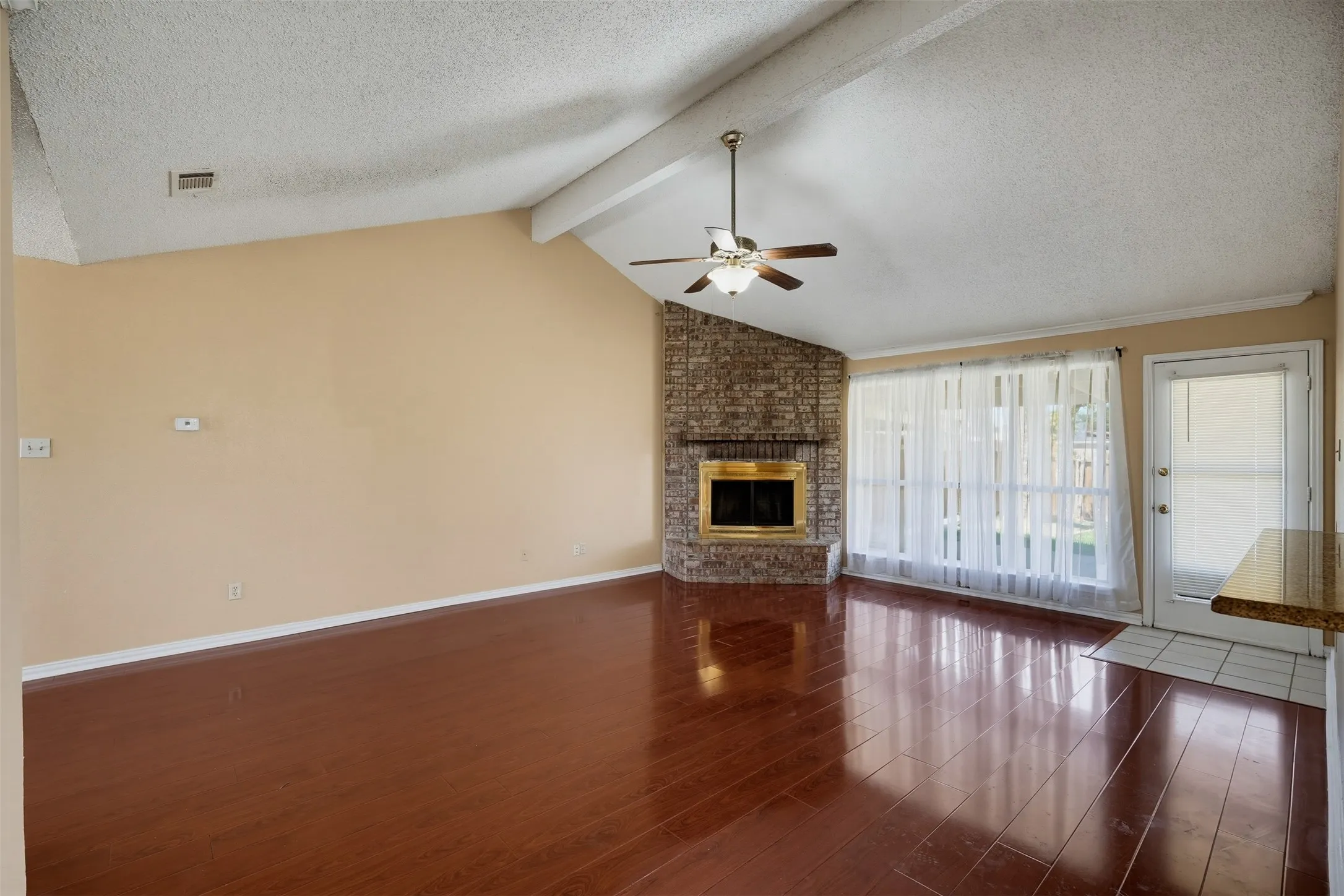 Vaulted ceiling with wood beam, wood-finish flooring, brick fireplace with brass firebox, ceiling fan with light fixture, and a white door with frosted glass​​‌​​​​‌​​‌‌​‌‌​​​‌‌​‌​‌​‌​​​‌​​ panel