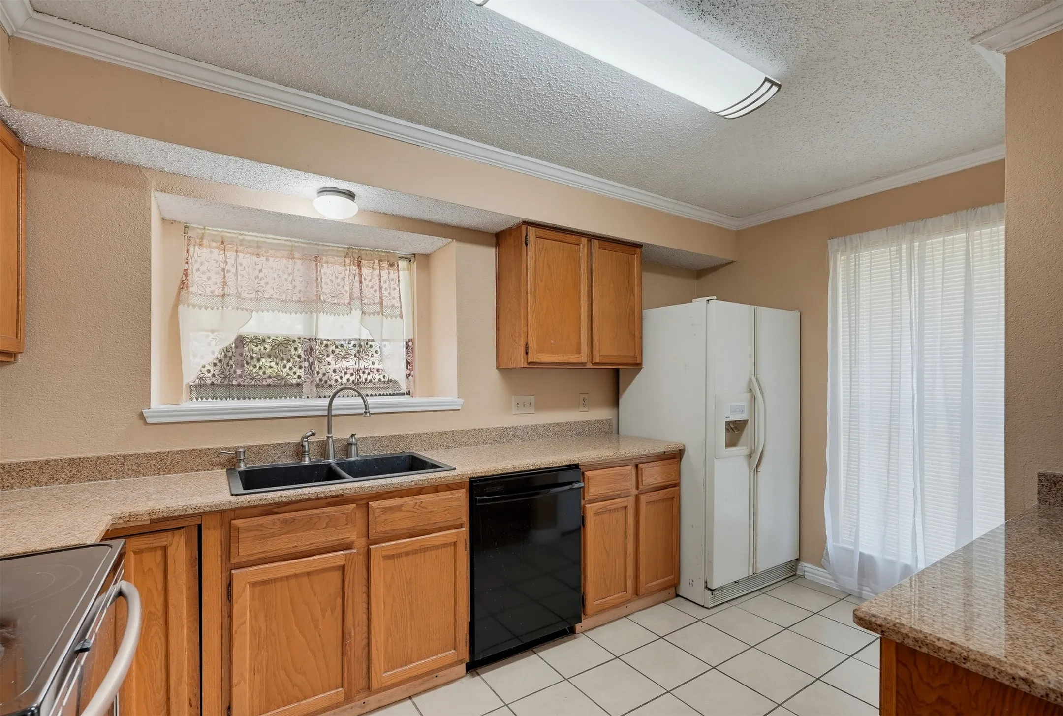 Kitchen with wood cabinetry, light-toned countertops, tile flooring, and a double basin sink
