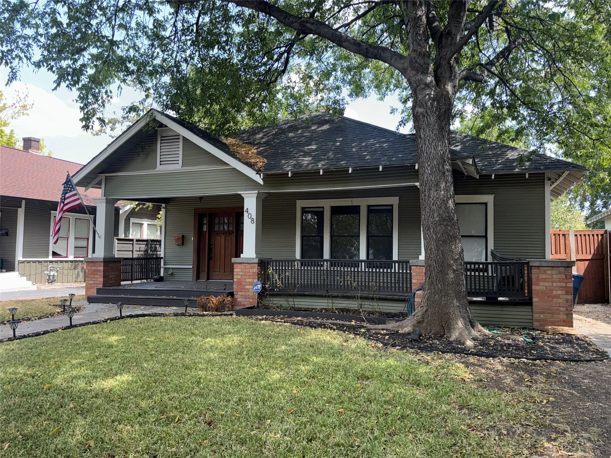 Craftsman-style exterior with a covered front porch, brick columns, wood-finish front door, and a mature front yard tree