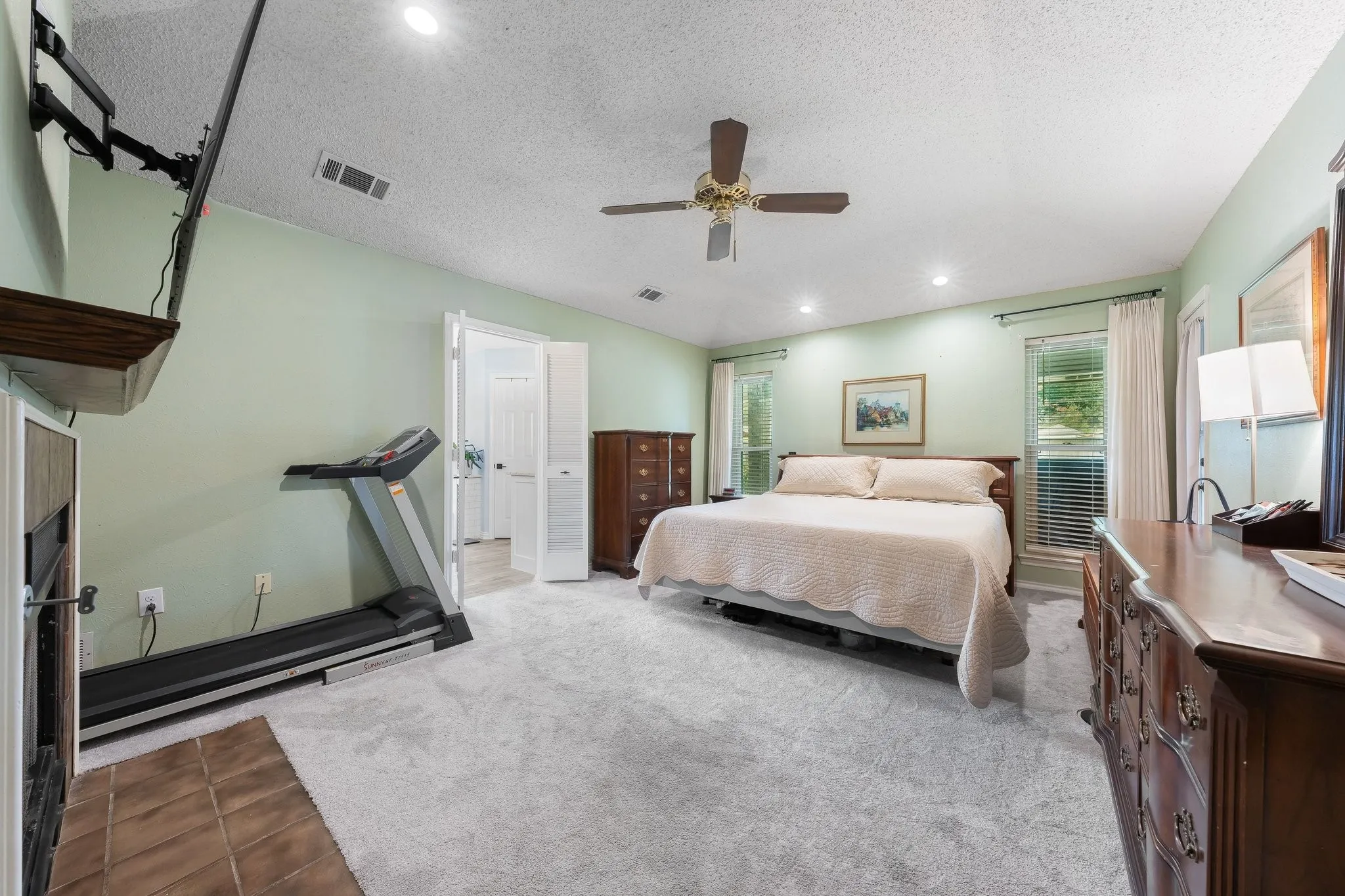 Spacious primary bedroom suite featuring soft sage-green walls, ceiling fan with integrated lighting, wood-finish flooring, ample natural light from multiple windows, and an attached ensuite bathroom