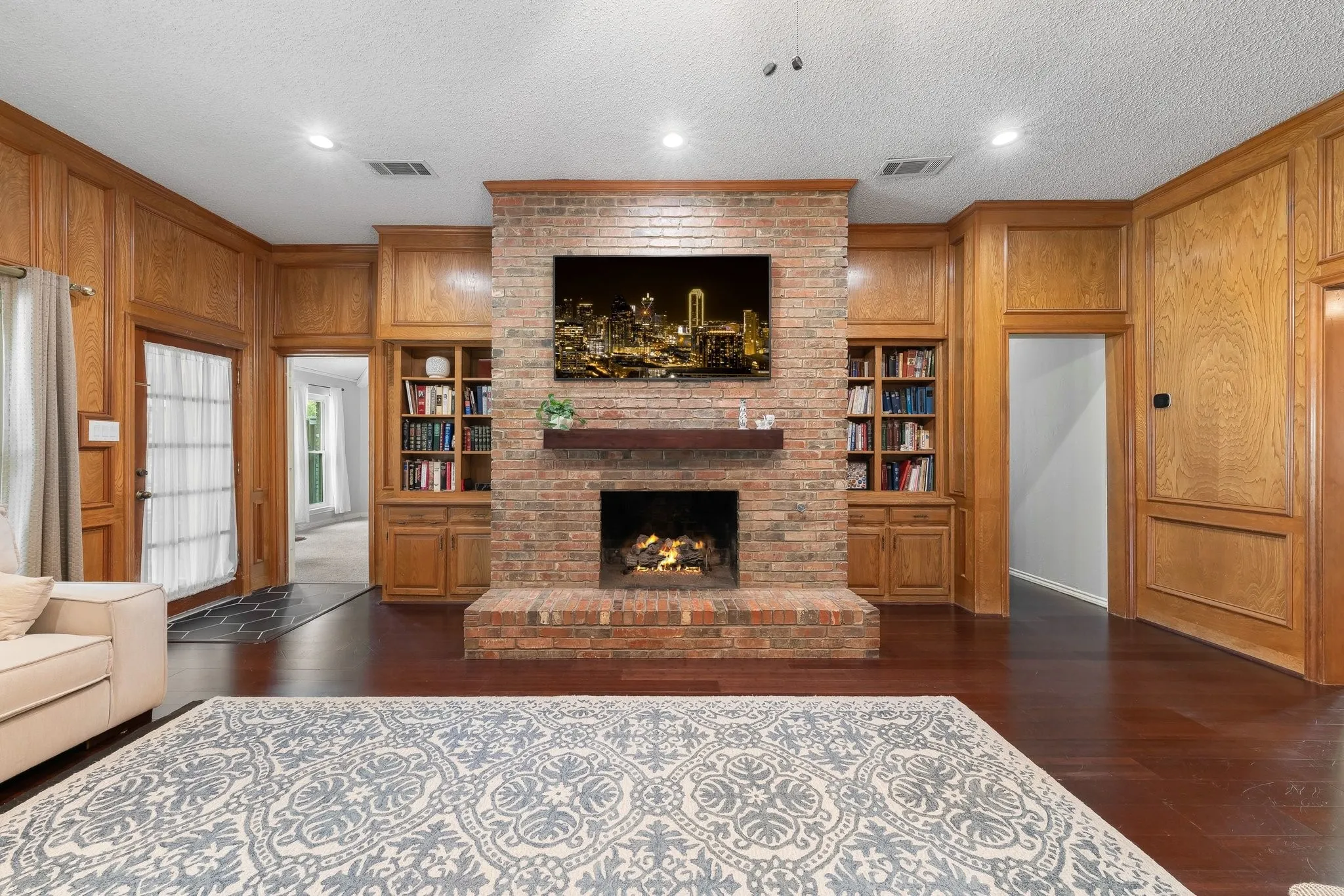 Brick fireplace with a wood mantel, rich wood-finish flooring, recessed lighting, and built-in cabinetry and shelving