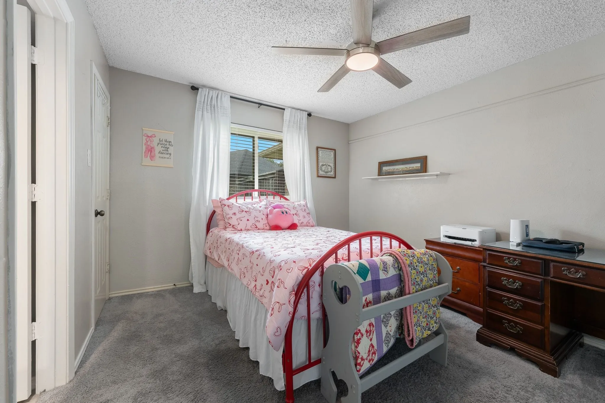Bright secondary bedroom featuring a red metal bed frame, carpet flooring, ceiling fan with integrated lighting, and wood-finish dresser with multiple drawers