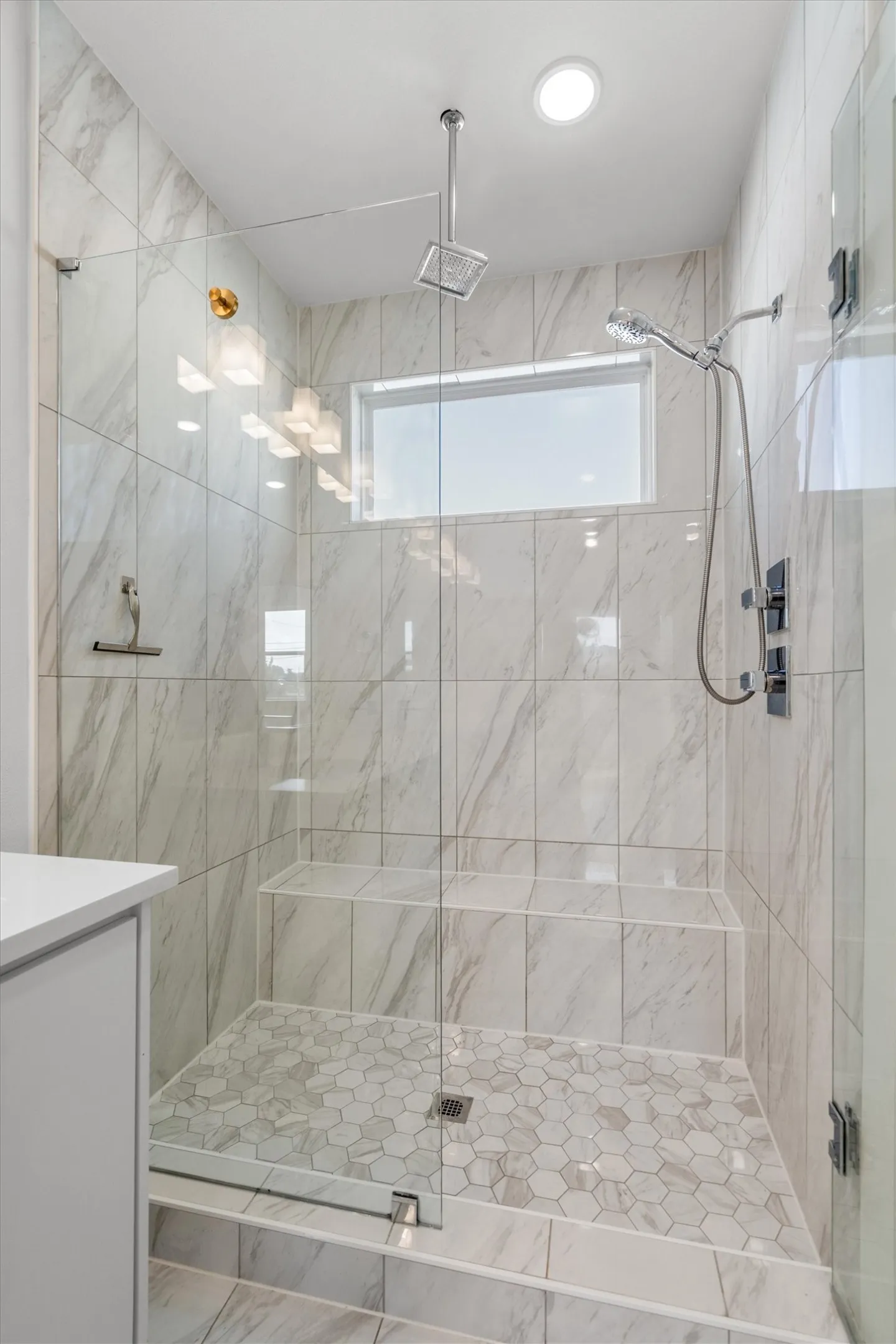 Walk-in shower with clear glass enclosure, tiled walls, hexagon-patterned floor, built-in bench, and a ceiling-mounted rainfall showerhead