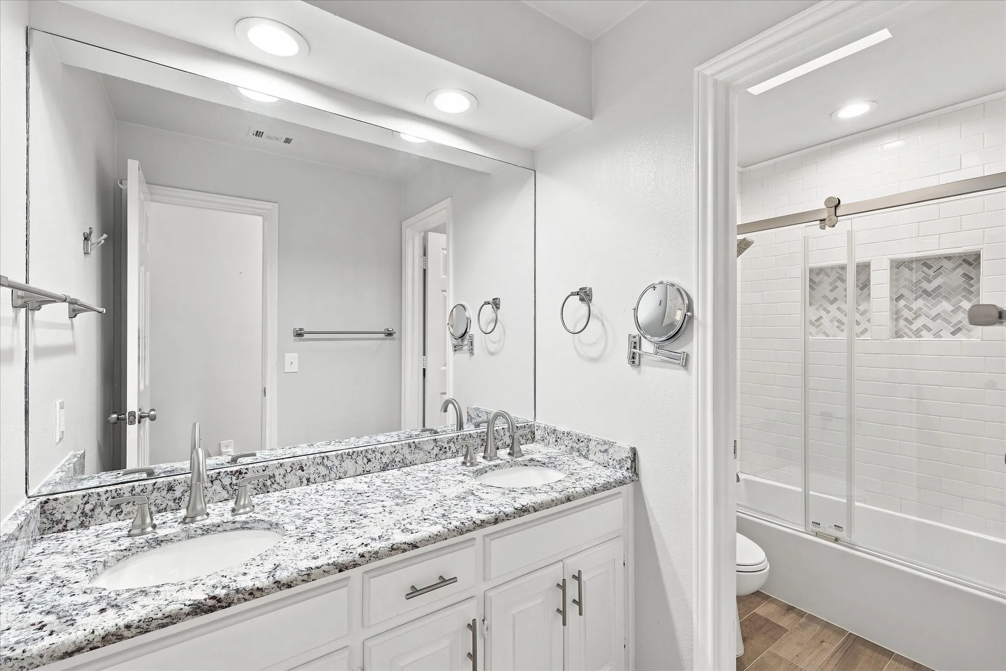 Dual vanity with granite countertop and undermount sinks, frameless mirror, recessed lighting, and a tub-shower combination with subway tile surround and decorative herringbone accent