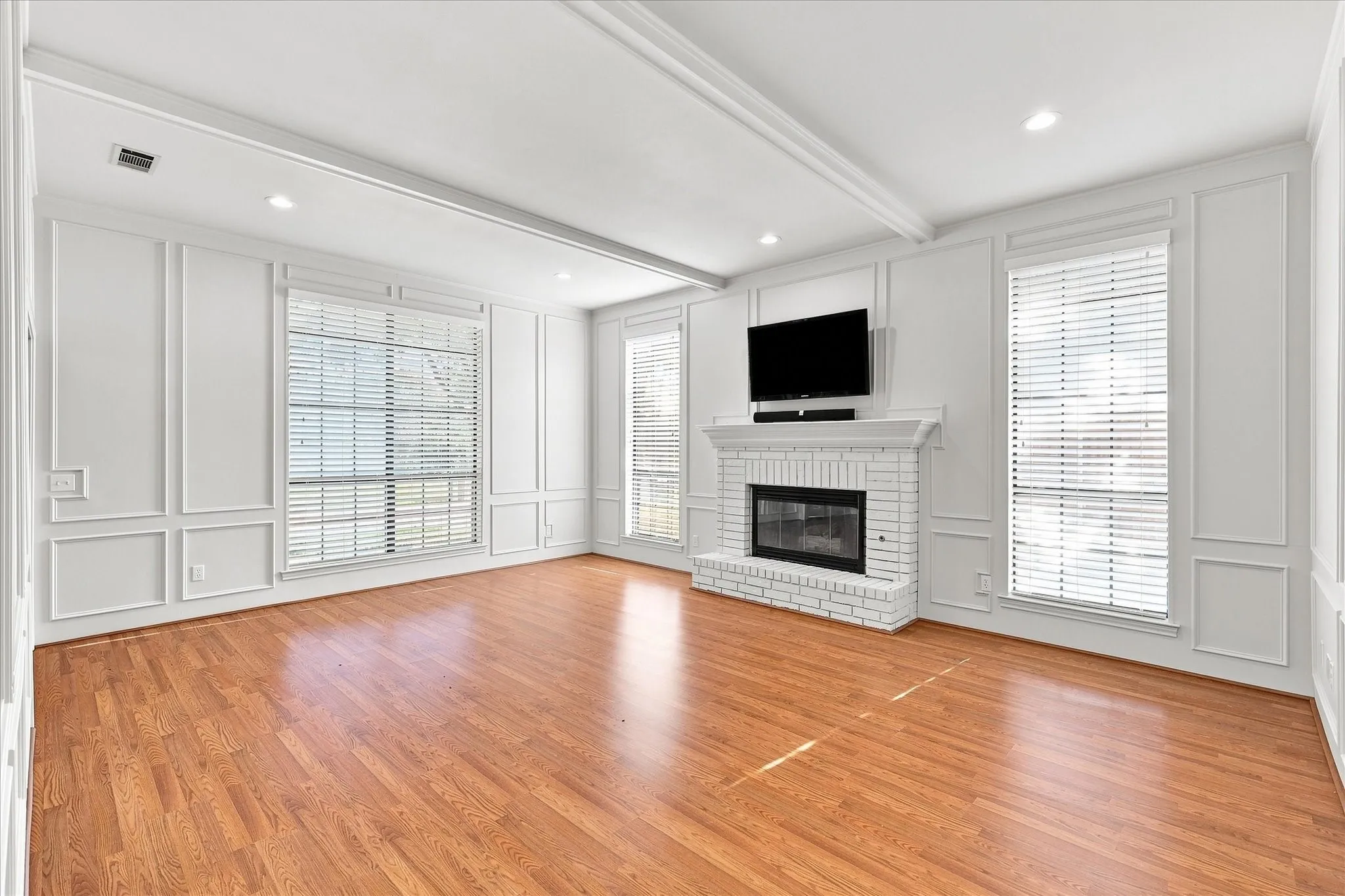 Hardwood flooring, painted brick fireplace, recessed lighting, white wall paneling, and large windows with blinds