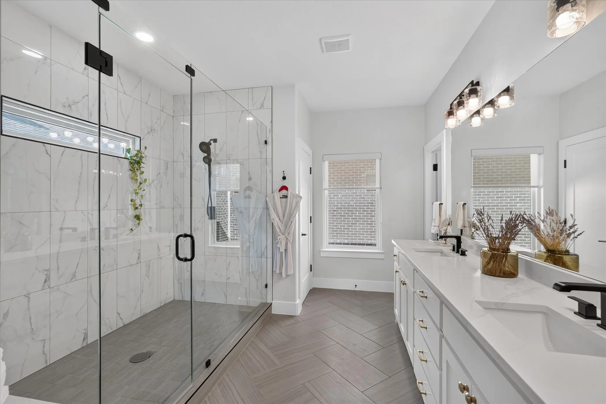 Spacious bathroom featuring a large walk-in shower with glass enclosure, chevron-patterned wood-finish flooring, a dual vanity with undermount sinks, and wall-mounted lighting fixtures