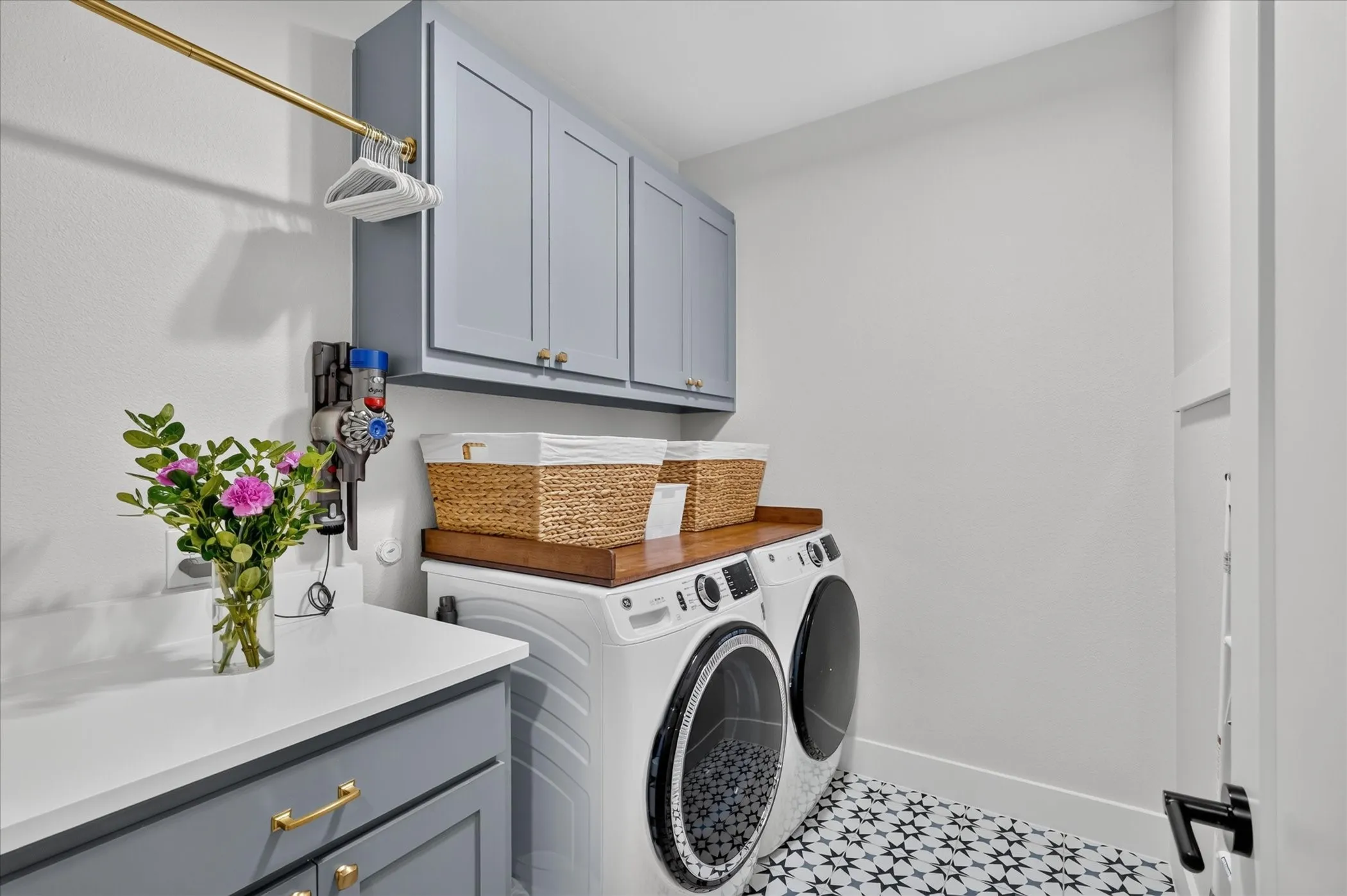 Laundry area featuring patterned floor tiles, light gray cabinetry with gold-toned hardware, a white countertop, and a wood-finish counter