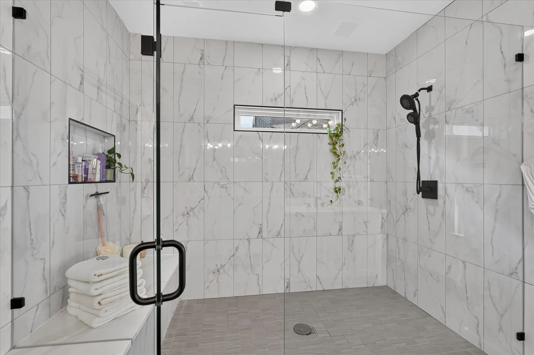 Spacious walk-in shower featuring glass enclosure, large format wall tiling, a built-in bench, and a recessed window