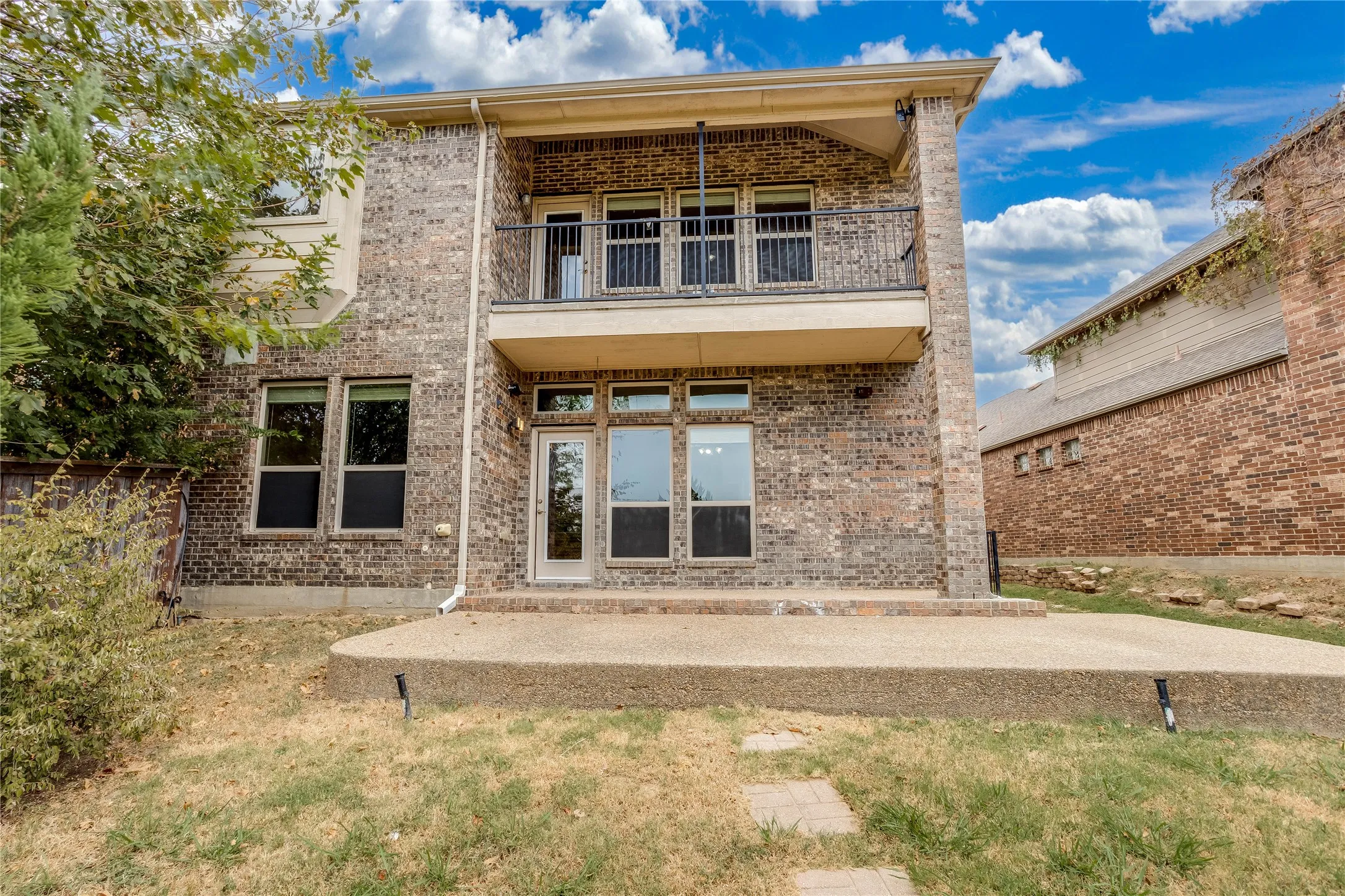 Brick exterior with multiple windows, a second-story balcony with metal railing, and a ground-level concrete patio