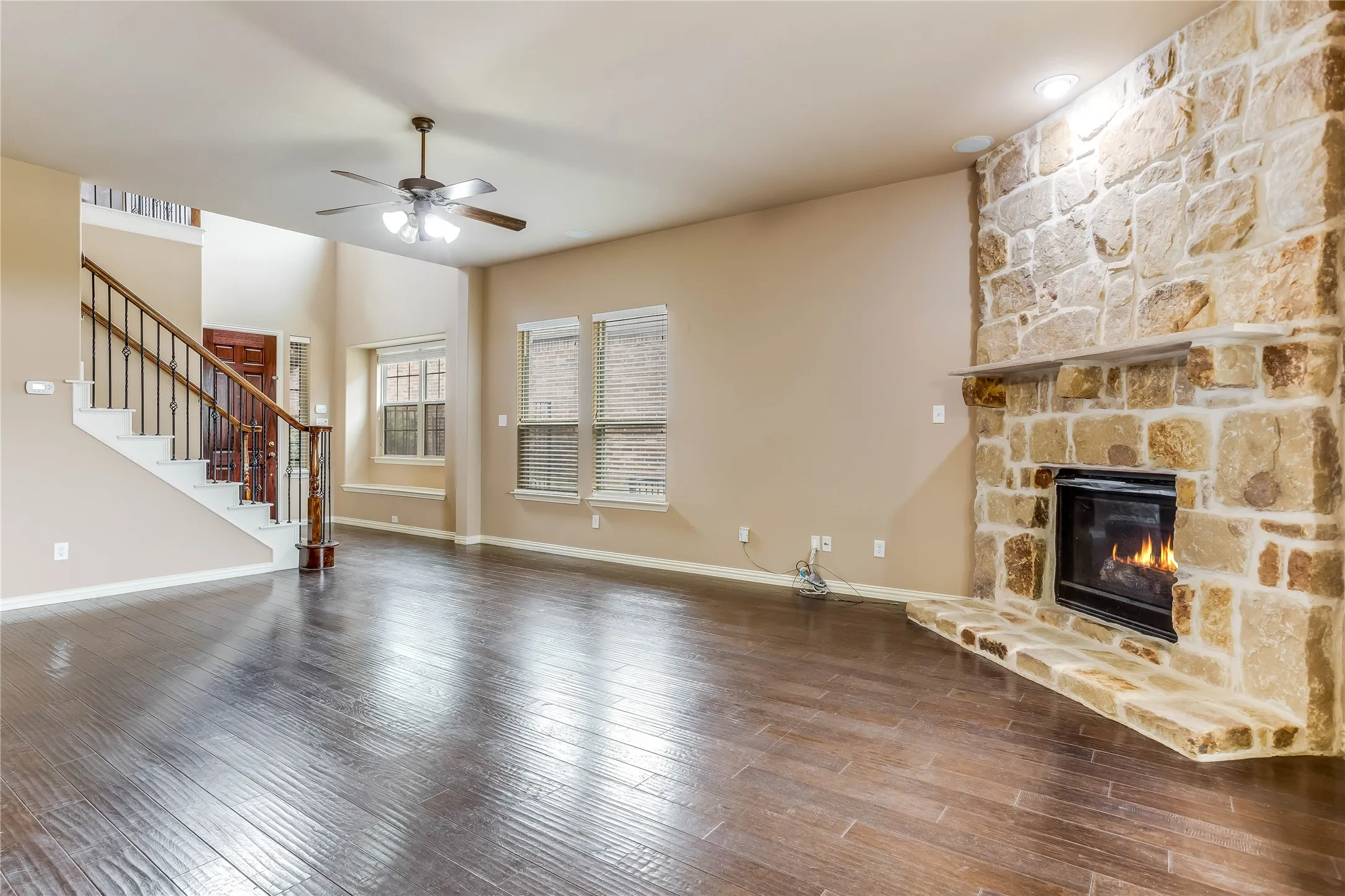 Stone-clad fireplace with hearth, dark wood-finish flooring, staircase with dark wood handrail and iron balusters, recessed lighting, and ceiling fan with integrated lighting