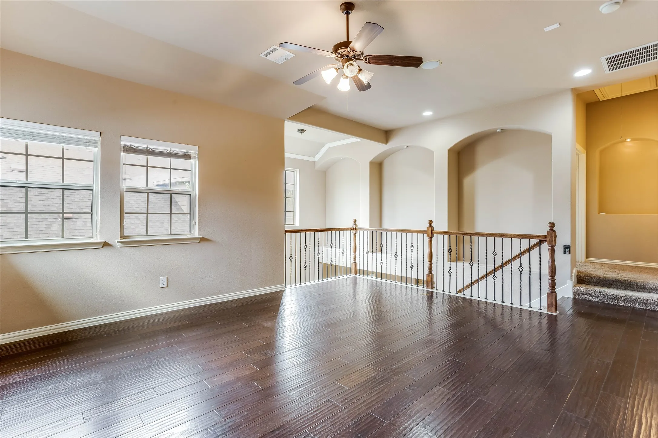 Dark wood-finish flooring, two large windows, a ceiling fan with integrated lighting, a wood and iron railing, and arched wall niches