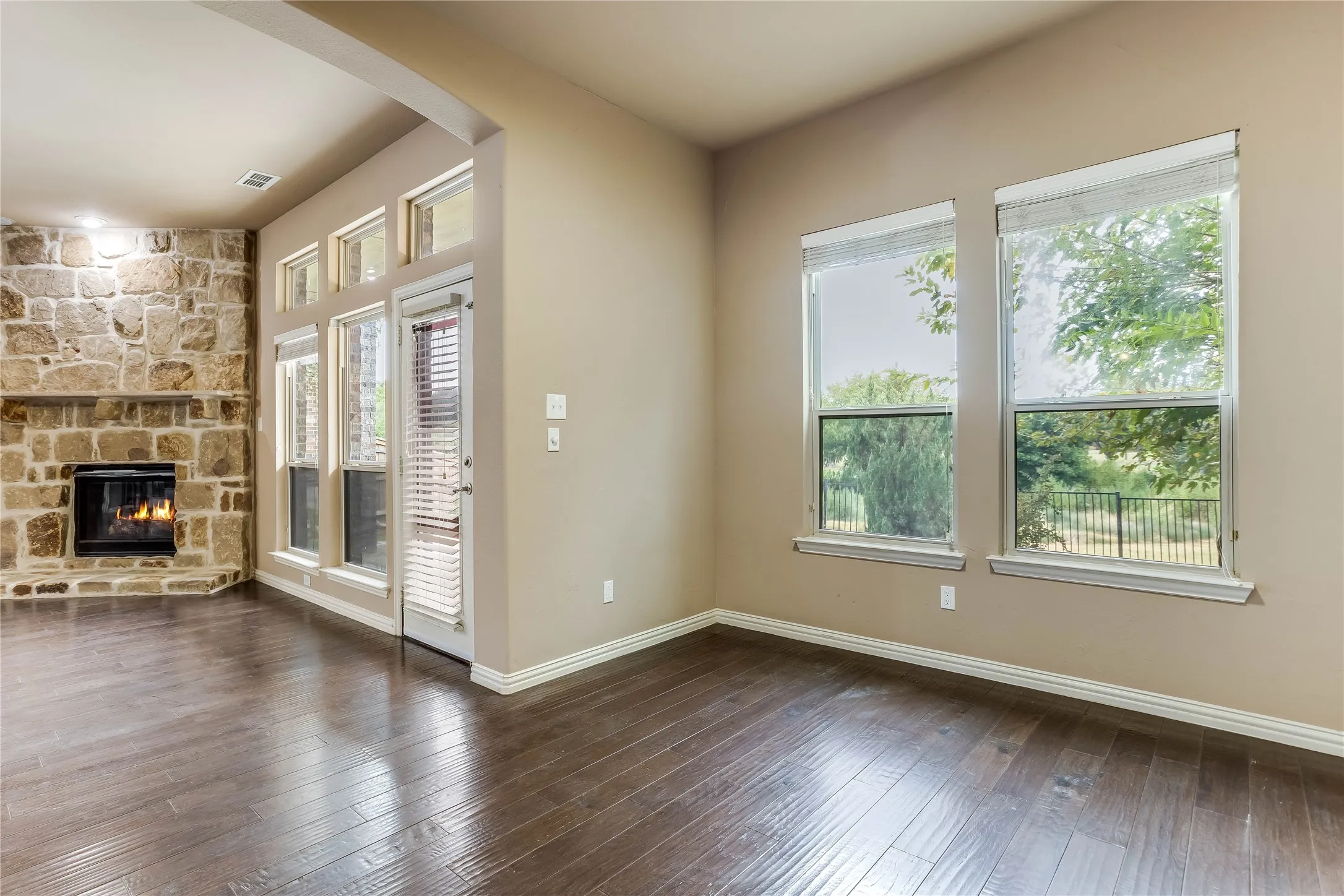 Stone-clad fireplace, wood-finish flooring, multiple large windows, transom window detailing, and an exterior door with integrated blinds