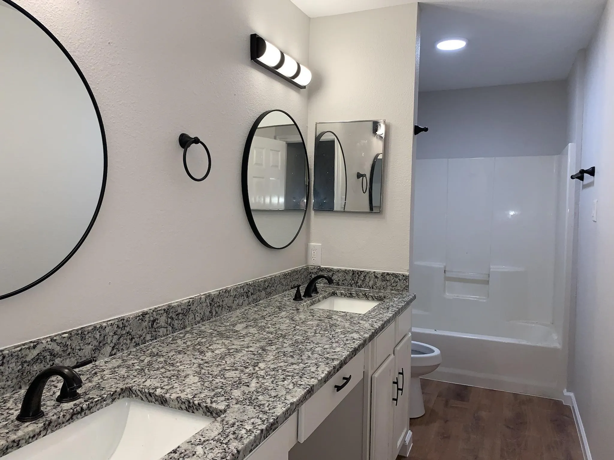 Dual undermount sinks with black fixtures, granite-look countertops, white cabinetry with black hardware, and wood-finish flooring