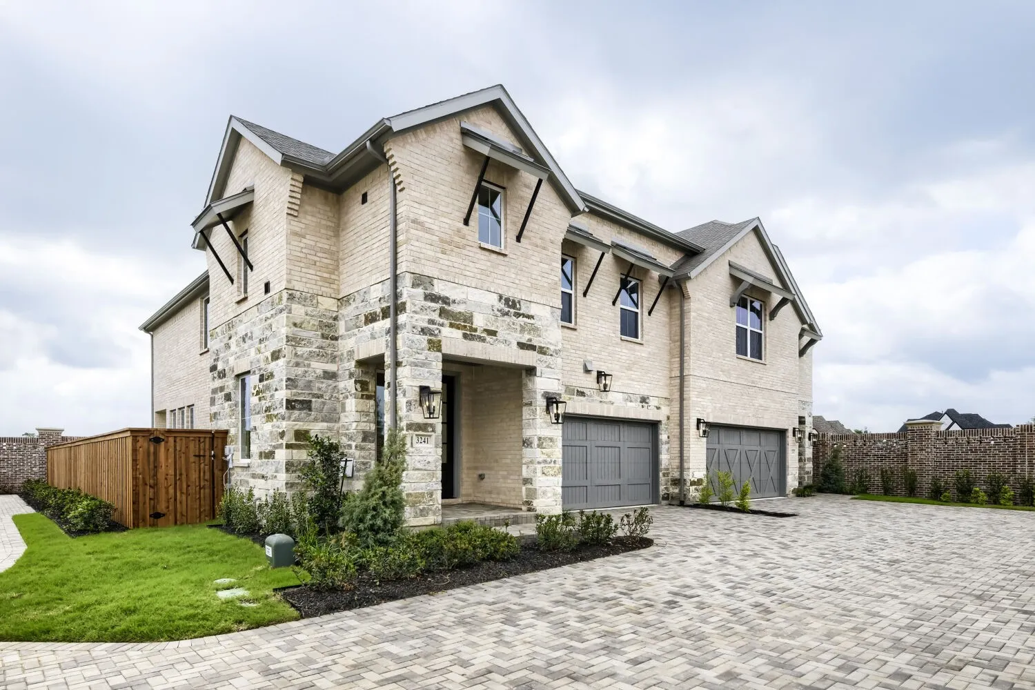 Two-story exterior with light brick and stone accents, two gray garage doors, a paver driveway, and a wooden privacy fence