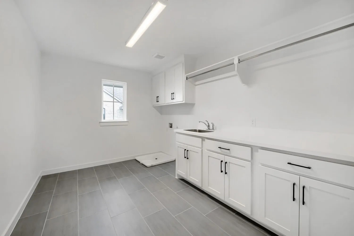 White cabinetry with black hardware, white countertop, stainless steel sink, grey tile flooring, and a single window