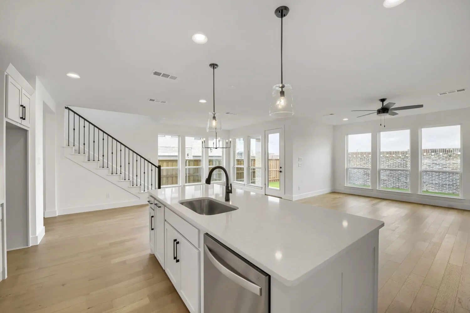 Open-concept interior featuring light wood-finish flooring, white cabinetry, a kitchen island with a built-in sink, stainless steel dishwasher, and contemporary pendant lighting