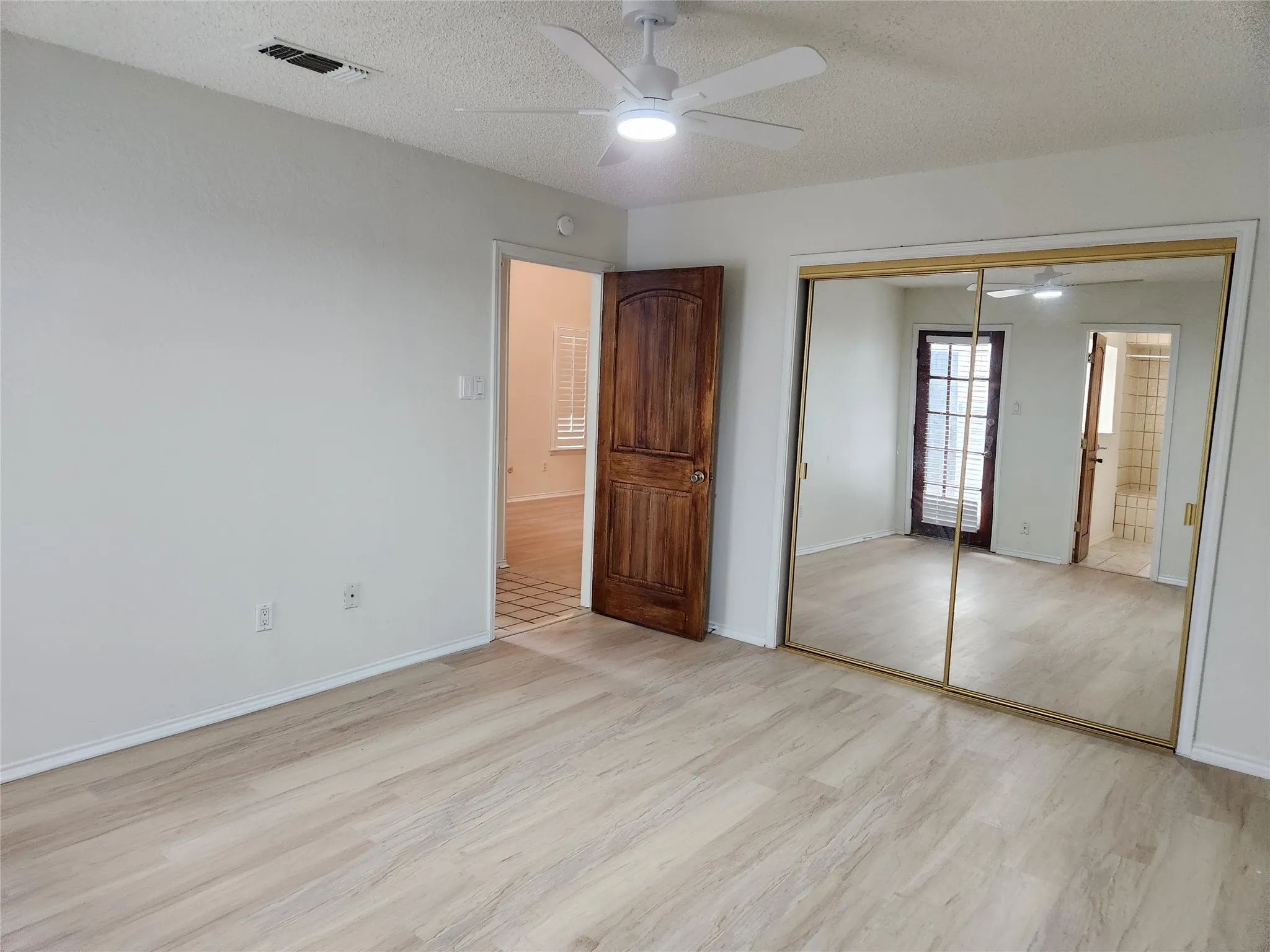 Light wood-finish flooring, white ceiling fan with integrated lighting, mirrored sliding closet doors, and a paneled wooden door