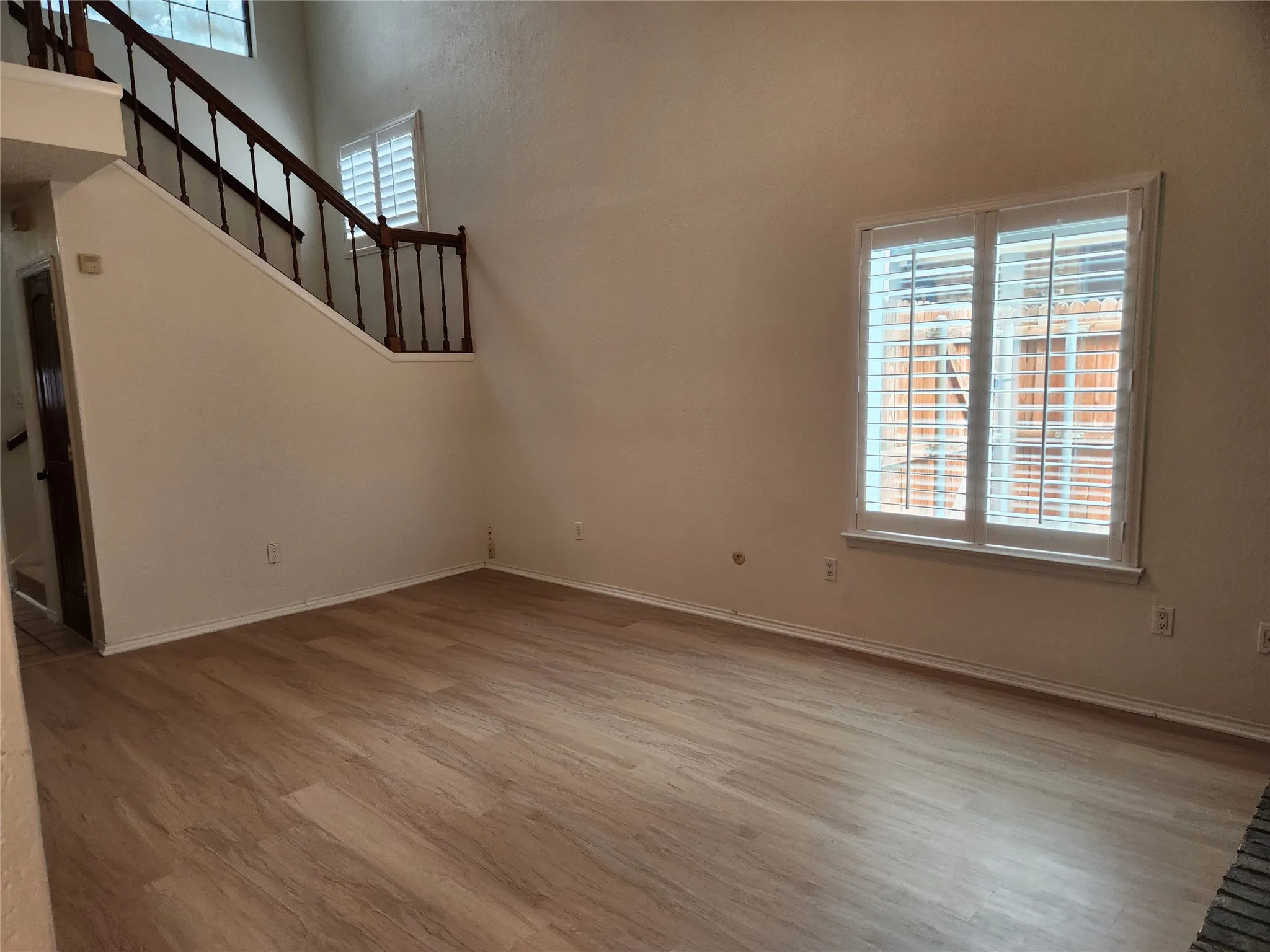 Spacious room featuring high ceilings, wood-finish flooring, a prominent staircase with dark wood railings, and windows with plantation shutters