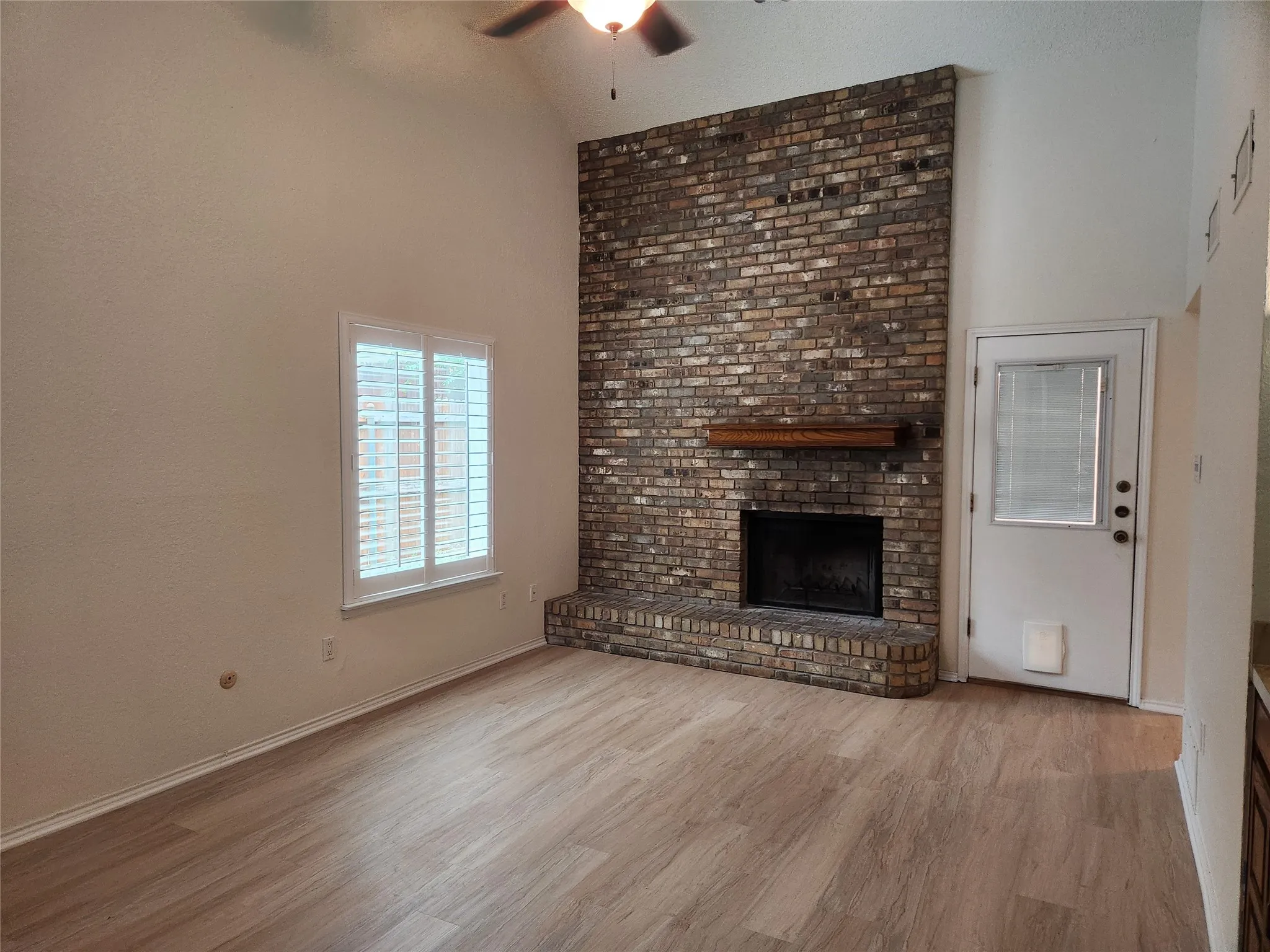 Vaulted ceiling with a ceiling fan, brick fireplace with wood mantel, wood-finish flooring, and window with plantation shutters