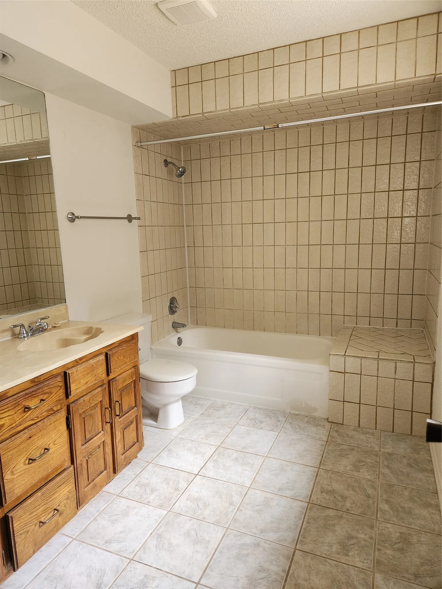 Spa-inspired bathroom featuring tile-lined shower surround with integrated showerhead and hand-held fixture, soaking tub, dual-sink vanity with wood cabinetry and brass hardware, and ceramic tile flooring throughout