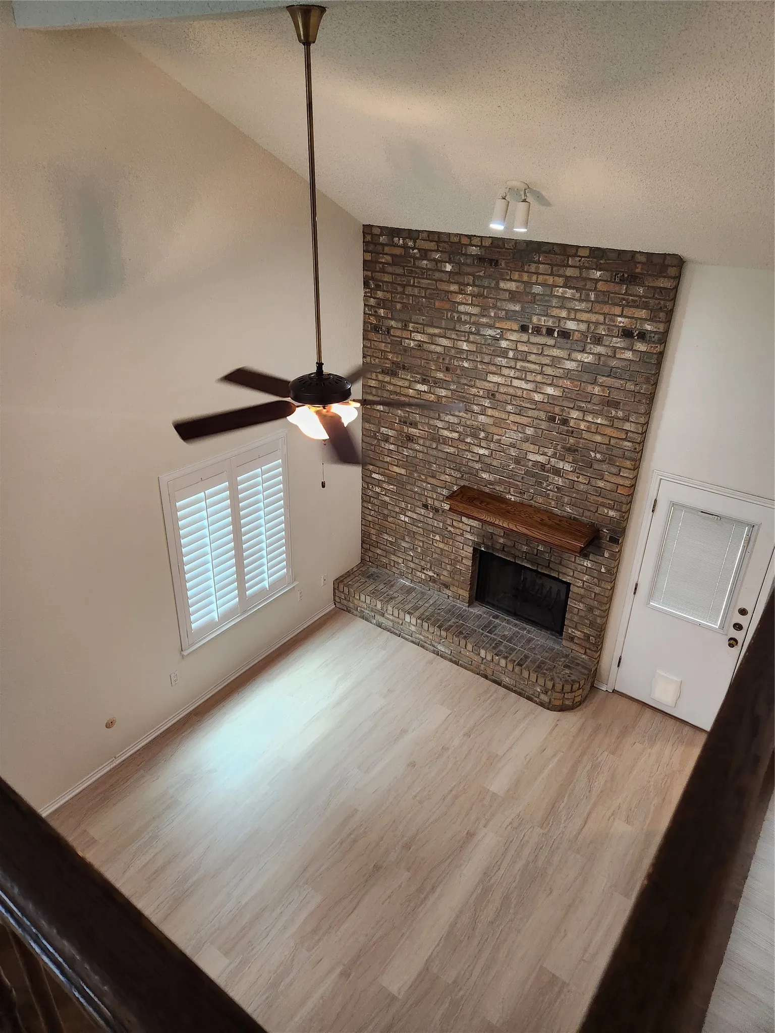 Vaulted ceilings with a ceiling fan, brick fireplace with a wood mantel, light wood-finish flooring, and a window with plantation shutters