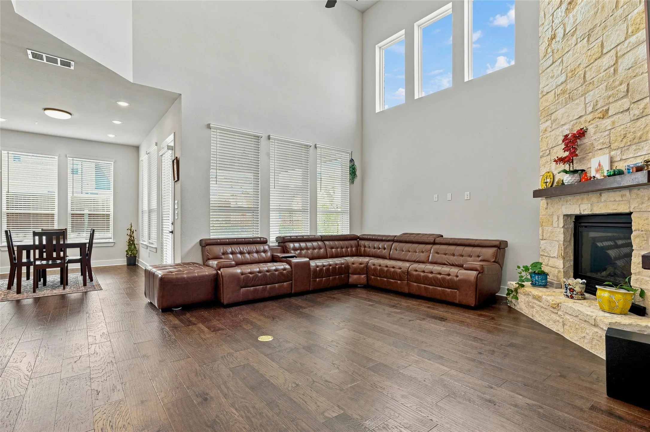 Spacious living area featuring high ceilings, wood-finish flooring, a stone-clad fireplace with a wood mantel, and multiple windows