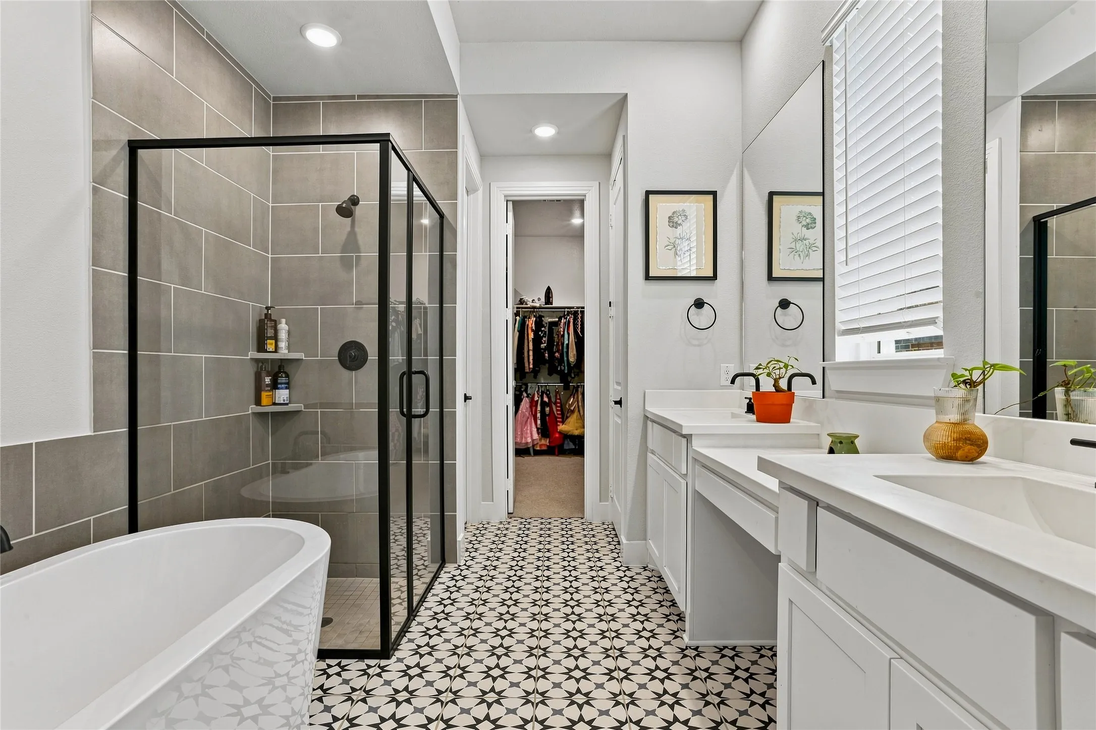 Patterned floor tile, dual vanities with white countertops, freestanding soaking tub, enclosed shower with gray tile surround, and a walk-in closet
